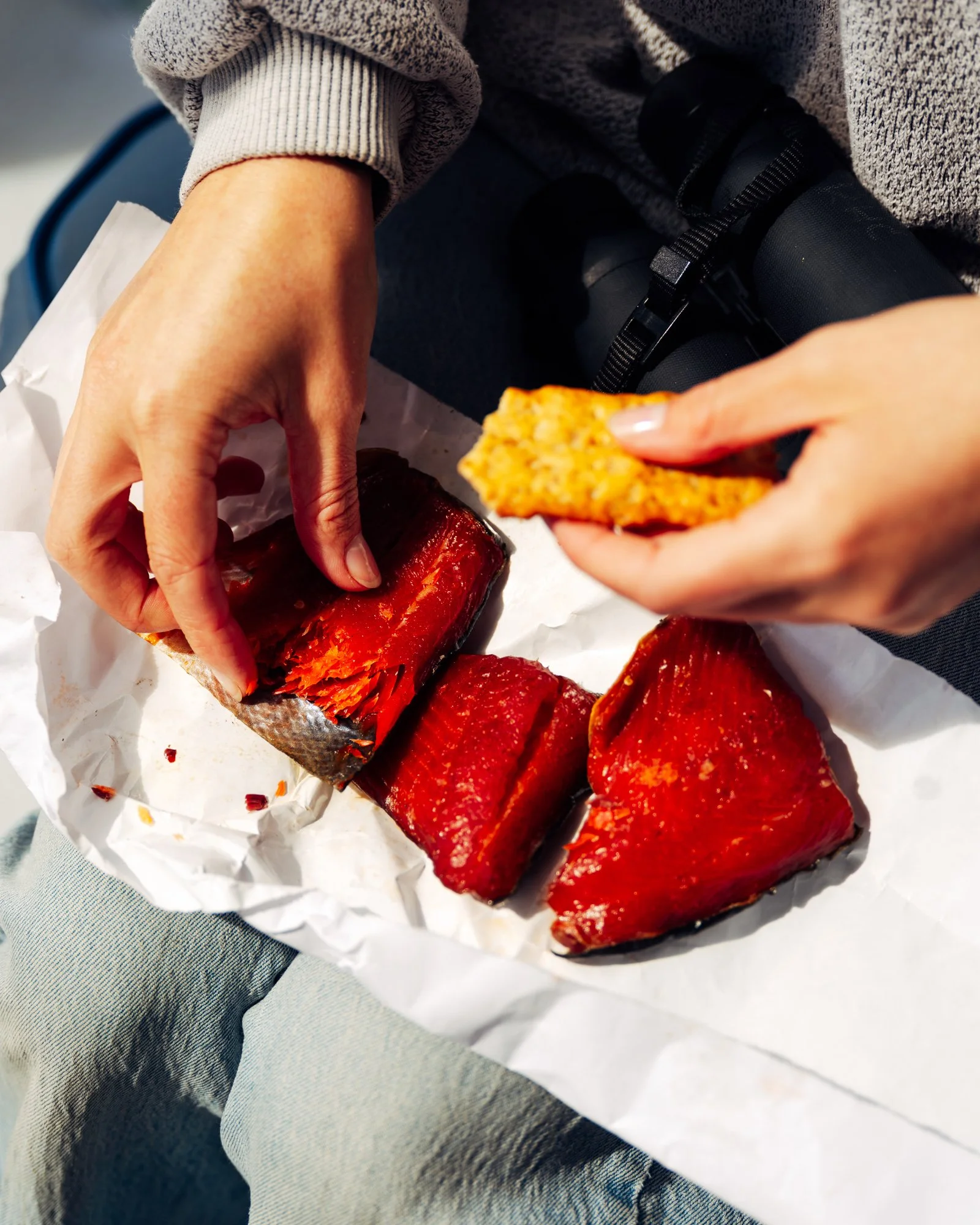 Close-up of a person's hands holding a piece of jerky over sliced beef jerky on white paper. The person is wearing a gray sweater and gray jeans.