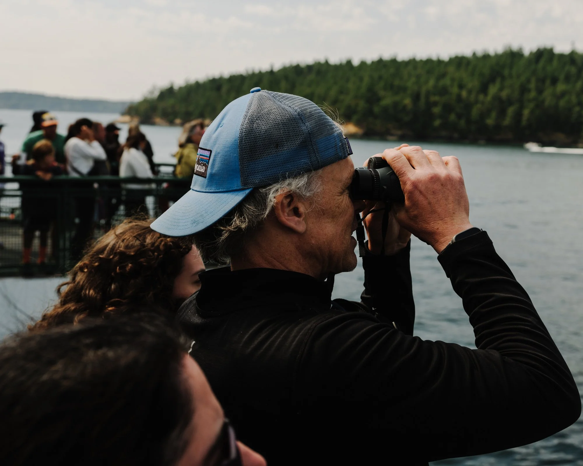 A person wearing a blue and gray Patagonia cap is looking through binoculars at a group of people on a boat or dock near a body of water, with a green forested area in the background.