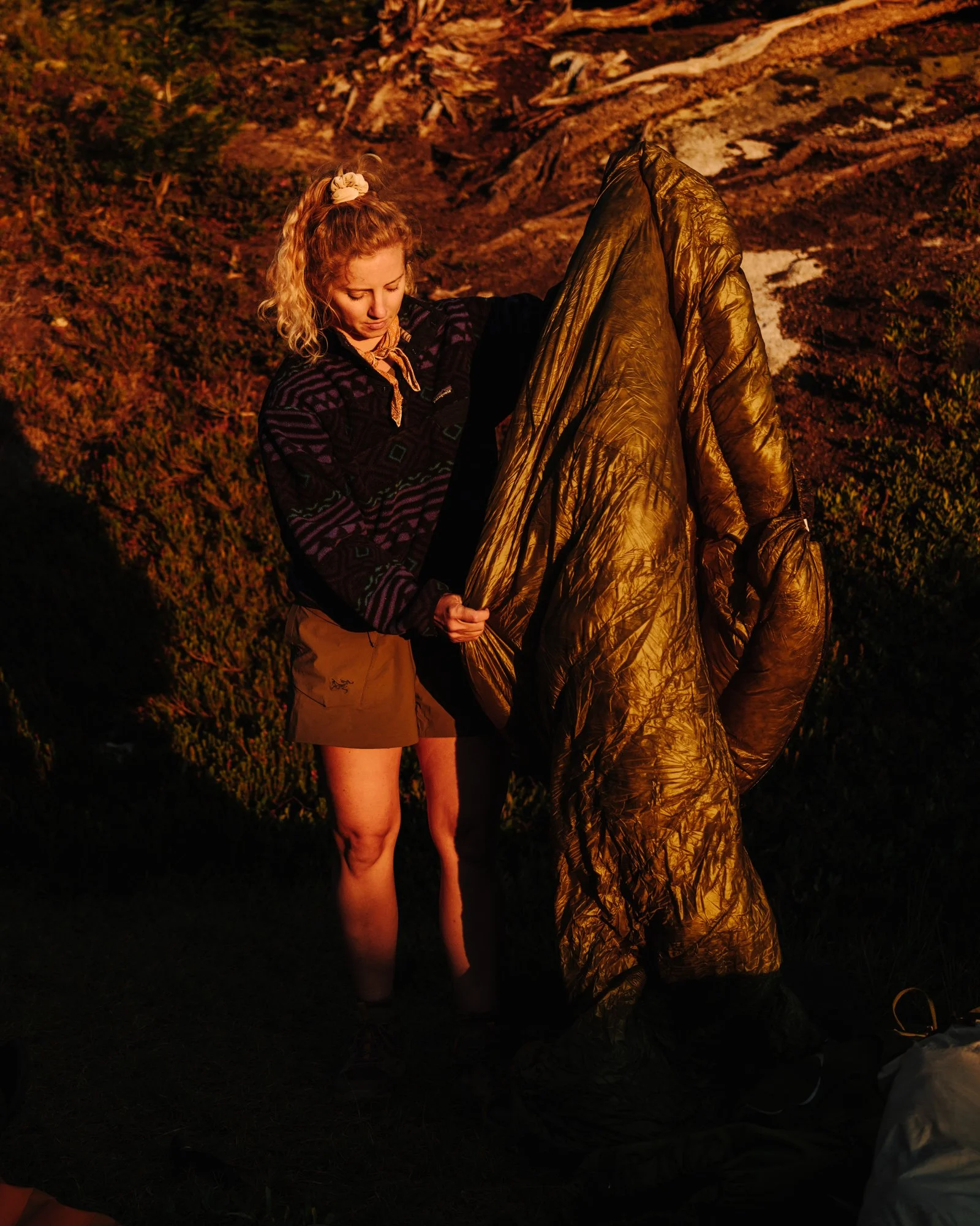 A woman with curly blonde hair tied up in a scrunchie, wearing a dark patterned jacket and tan shorts, stands outdoors at sunset. She is holding a shiny gold sleeping bag or blanket and looking down with a focused expression. The background features 