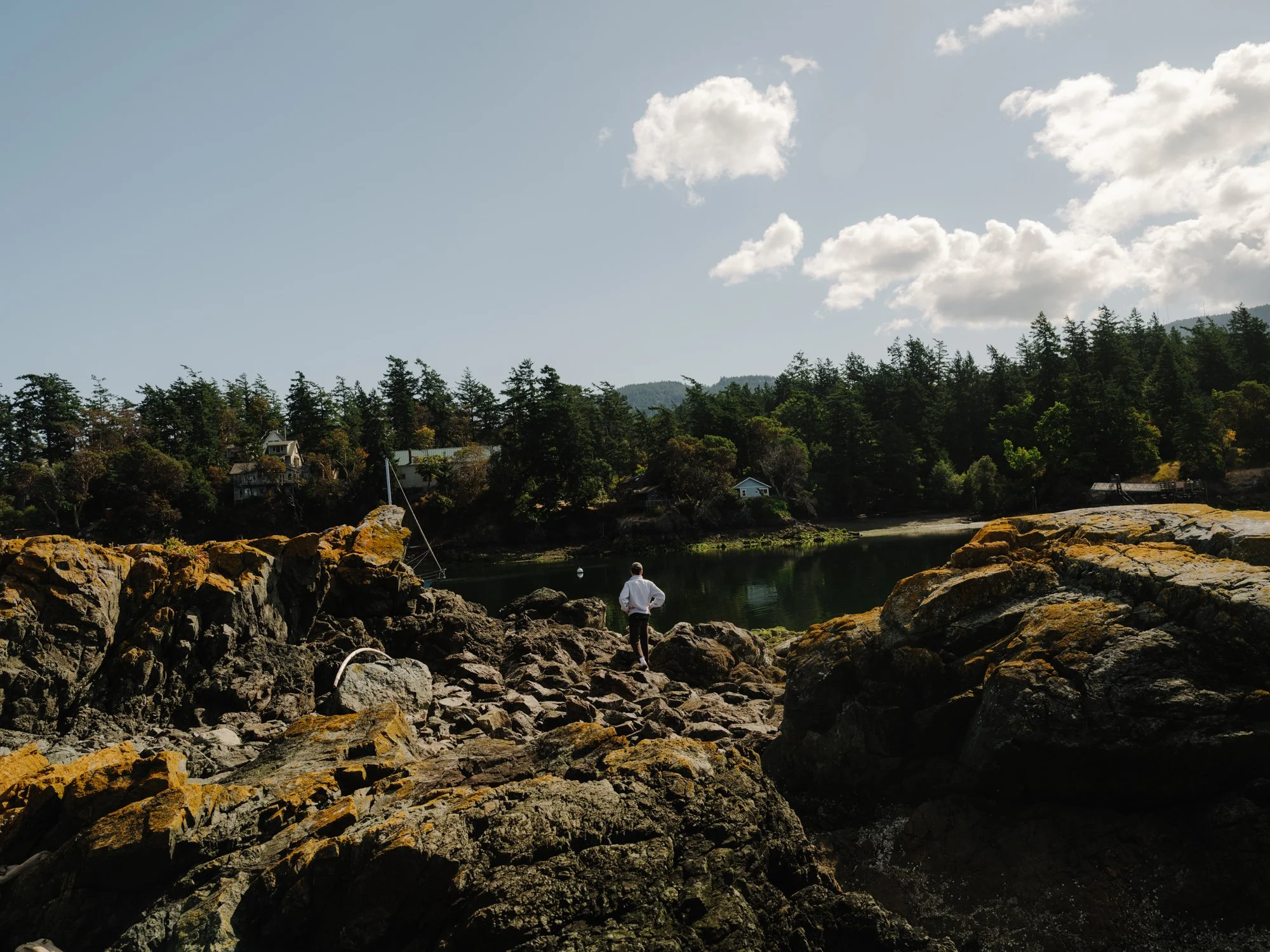 A man standing on rocks by a river or lake, with a forested hillside and houses in the background, under a partly cloudy sky.