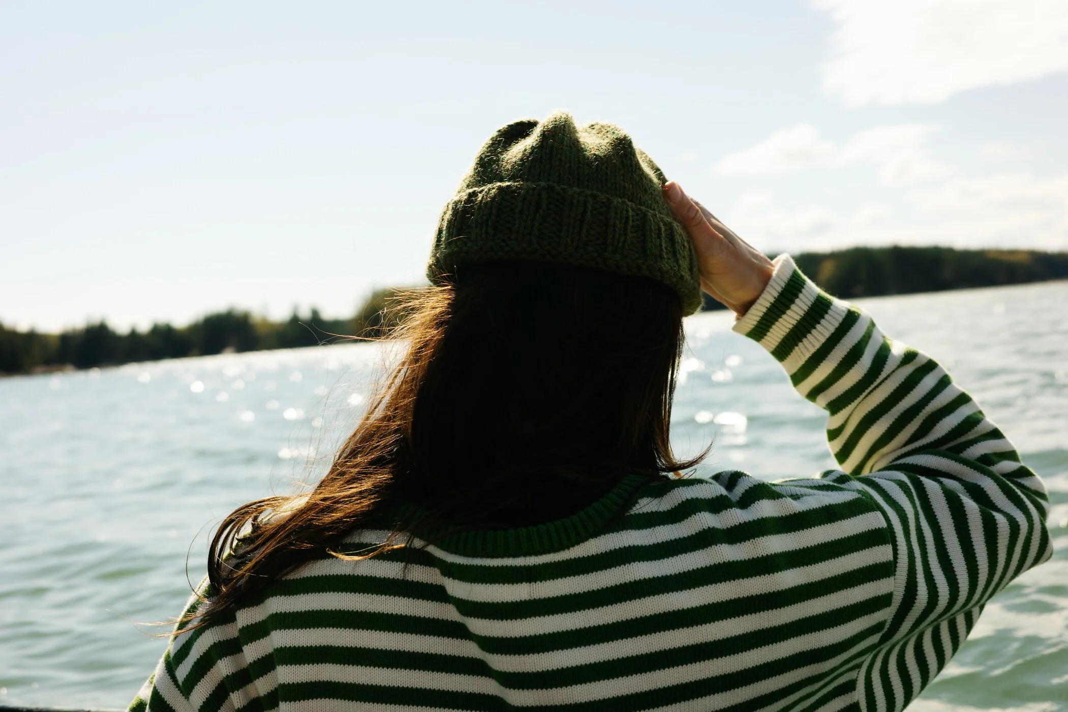 A person with long hair wearing a green knit beanie and a green and white striped sweater, standing outdoors near water with trees in the background, holding the beanie on their head.