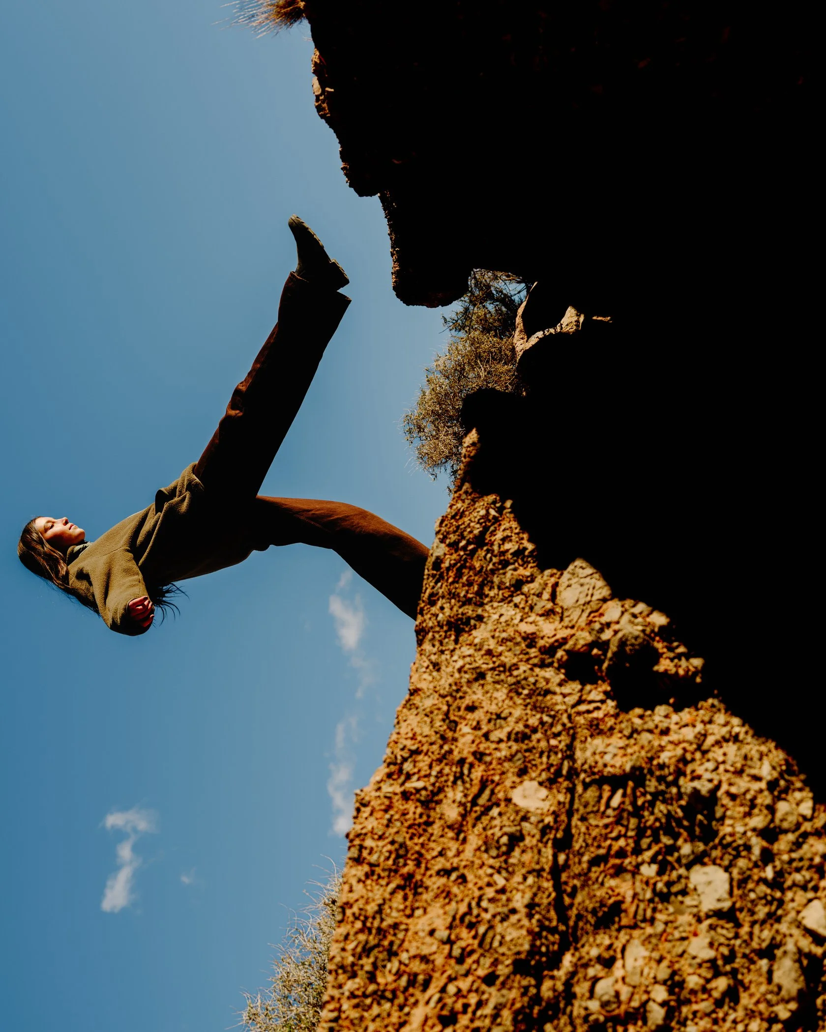 A person is laying on a rocky surface outdoors, with a clear blue sky behind them. They are wearing a green jacket, dark pants, and a hat, and are reaching towards a rock formation.