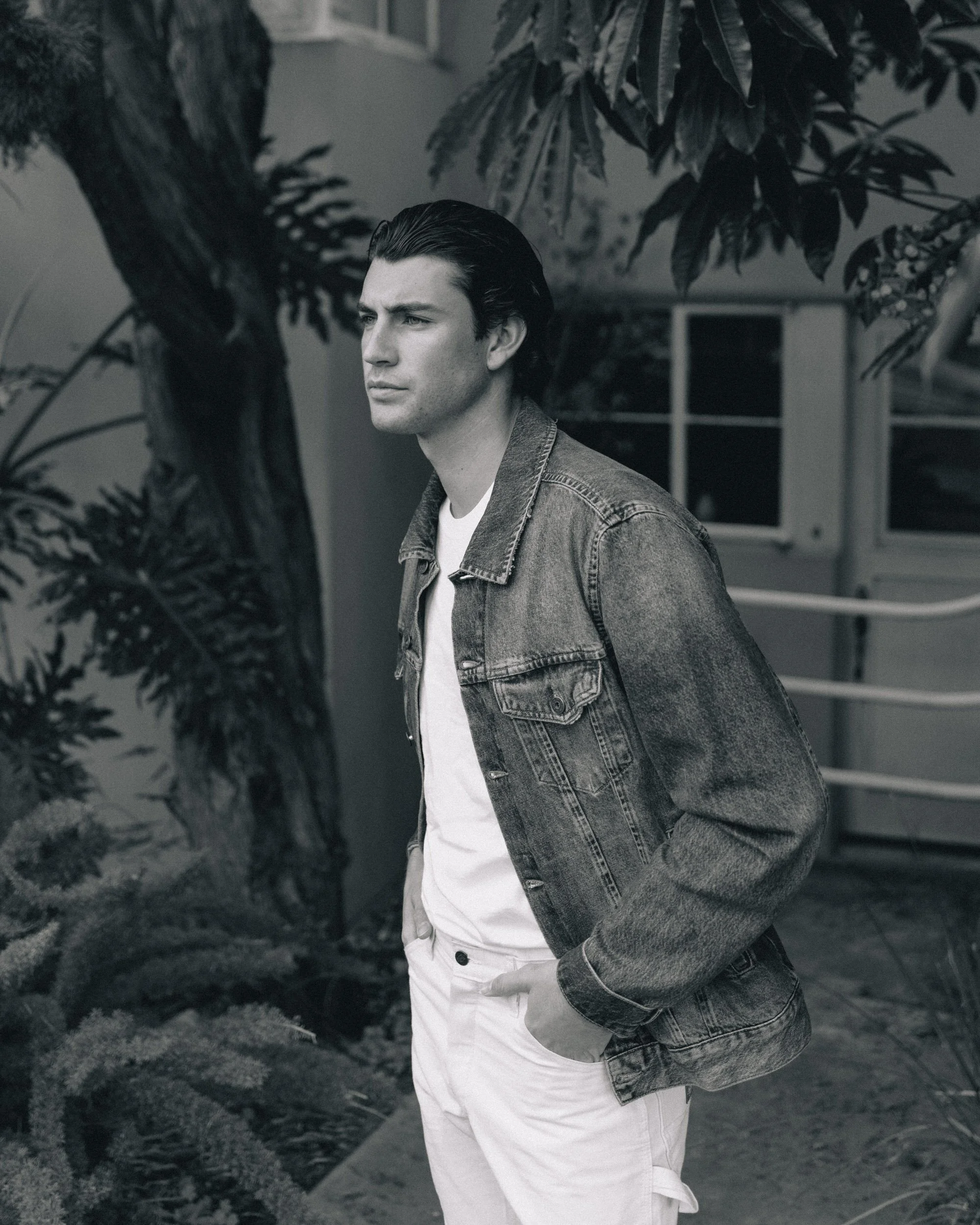 A young man with dark, slicked-back hair standing outdoors near a tree with large leaves. He wears a denim jacket over a white t-shirt and white pants, with his hand in his pocket. The background includes a house with a window and a staircase.