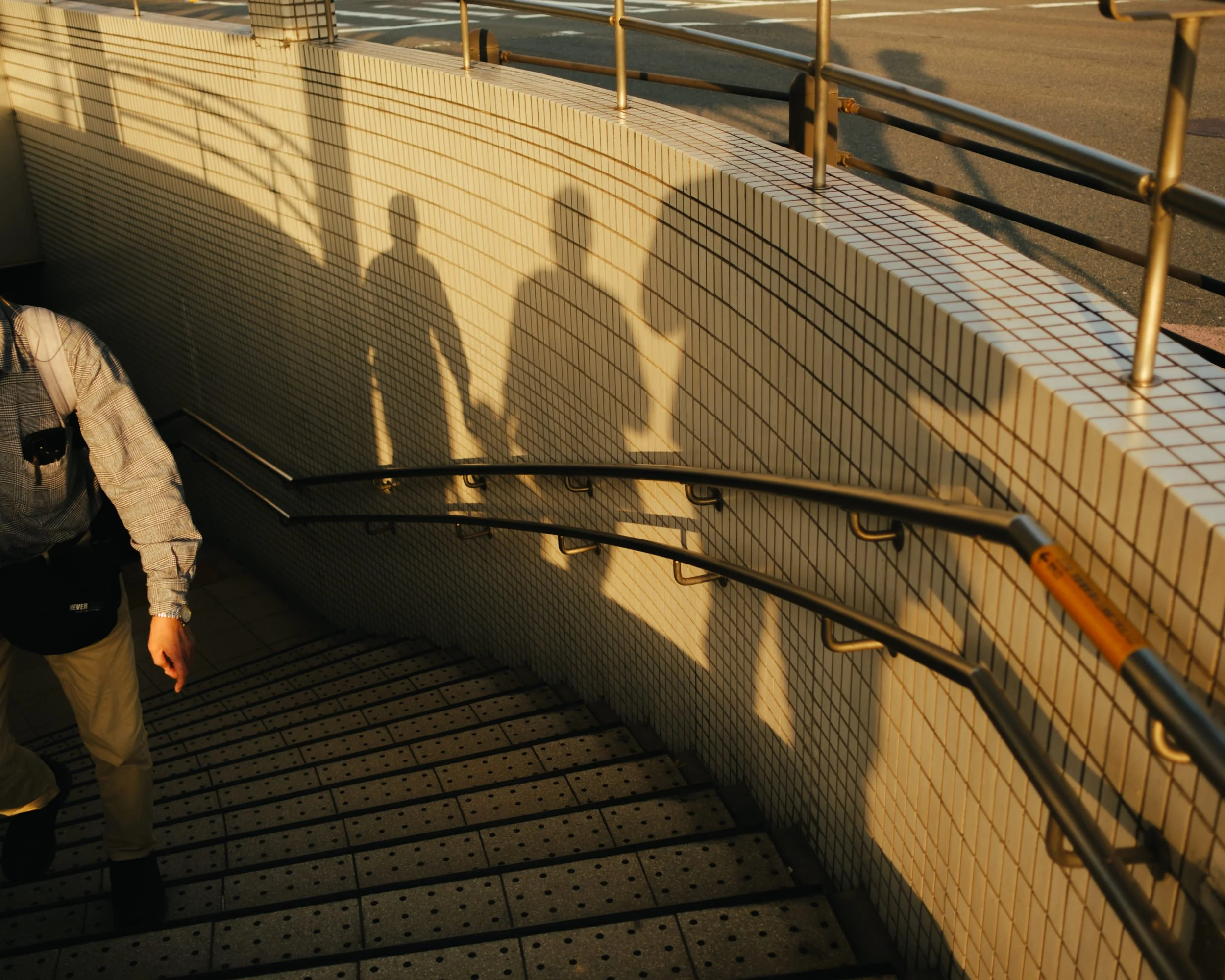 Shadow of two people walking along a tiled wall on a staircase leading outside, with a handrail on the right side. The sunlight creates long shadows.
