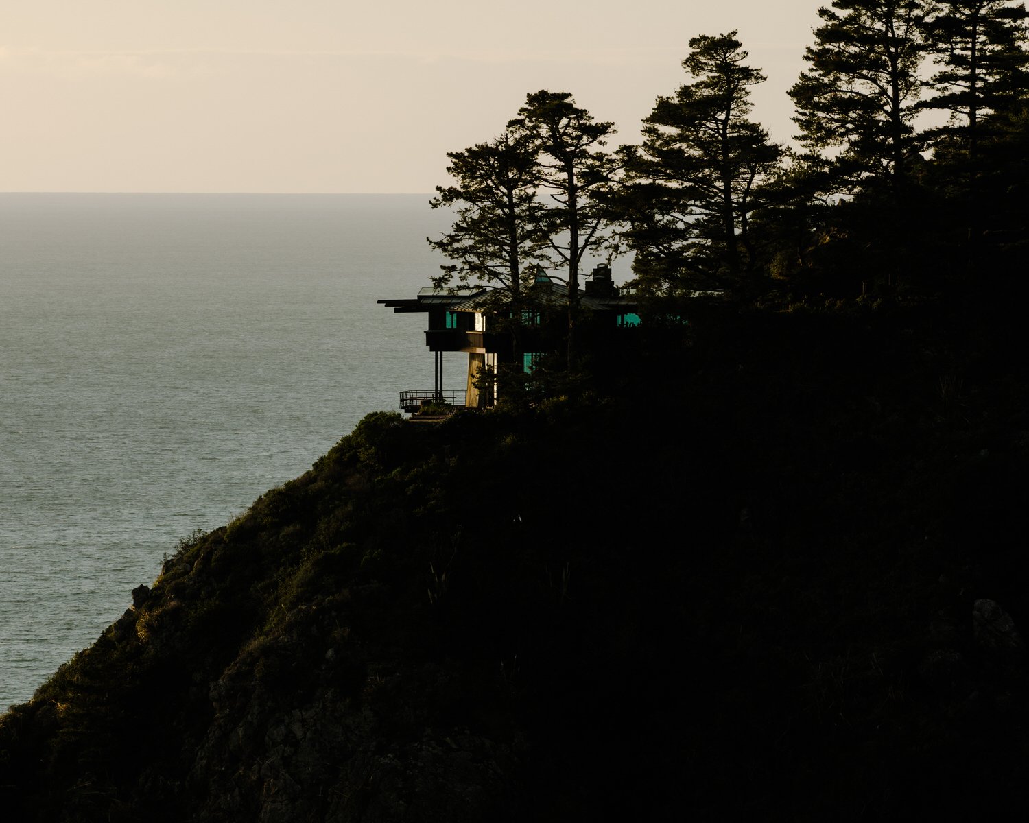 A house overlooking the ocean on a cliff, surrounded by tall trees at sunset