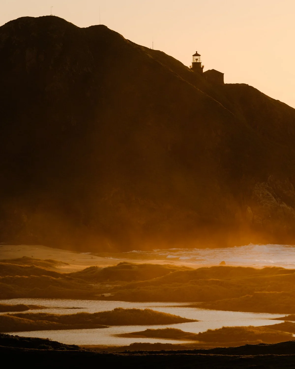 Sunset over a beach with waves crashing and a lighthouse on a hill in the distance.