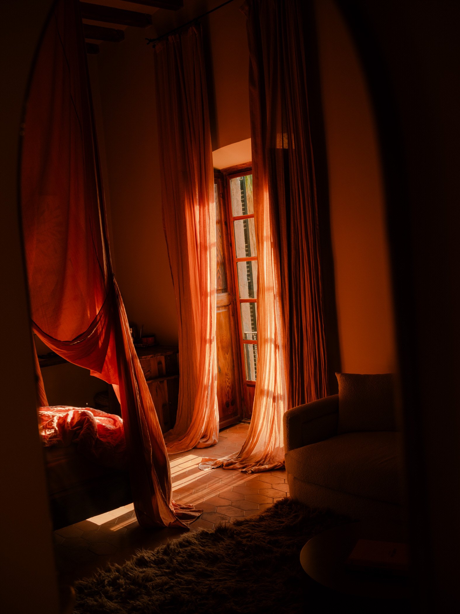 Sunlight streams through open window curtains into a cozy room with a bed and armchair, creating a warm, inviting atmosphere.