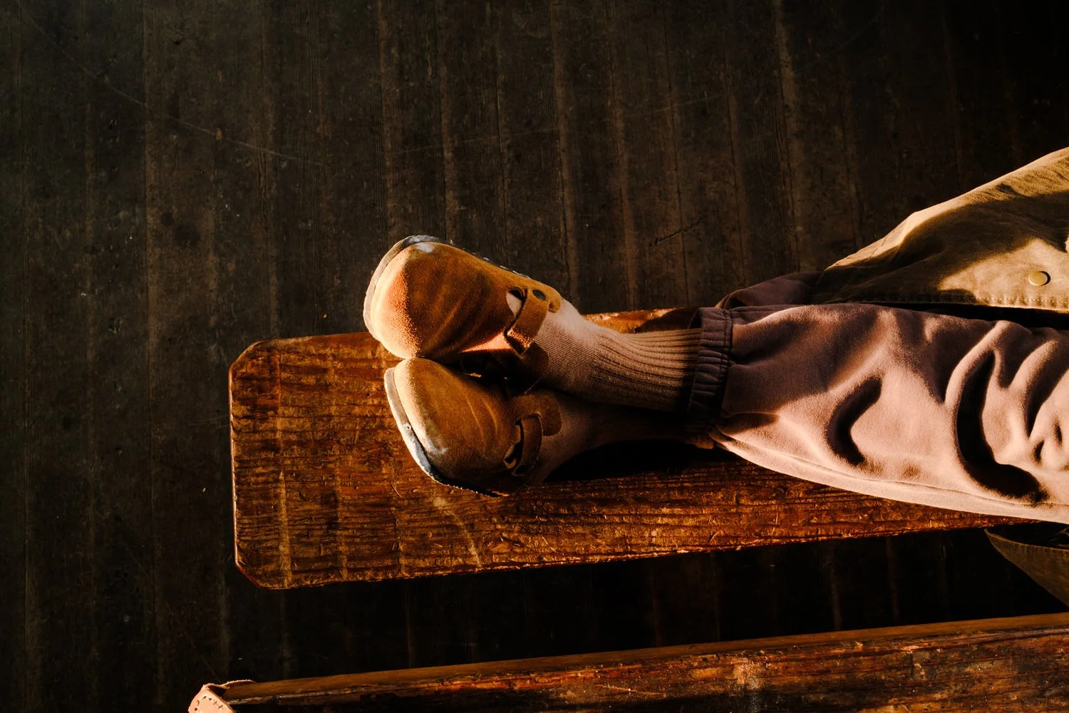 A person sitting on a wooden bench wearing beige shoes, gray socks, and gray sweatpants with a tan jacket.