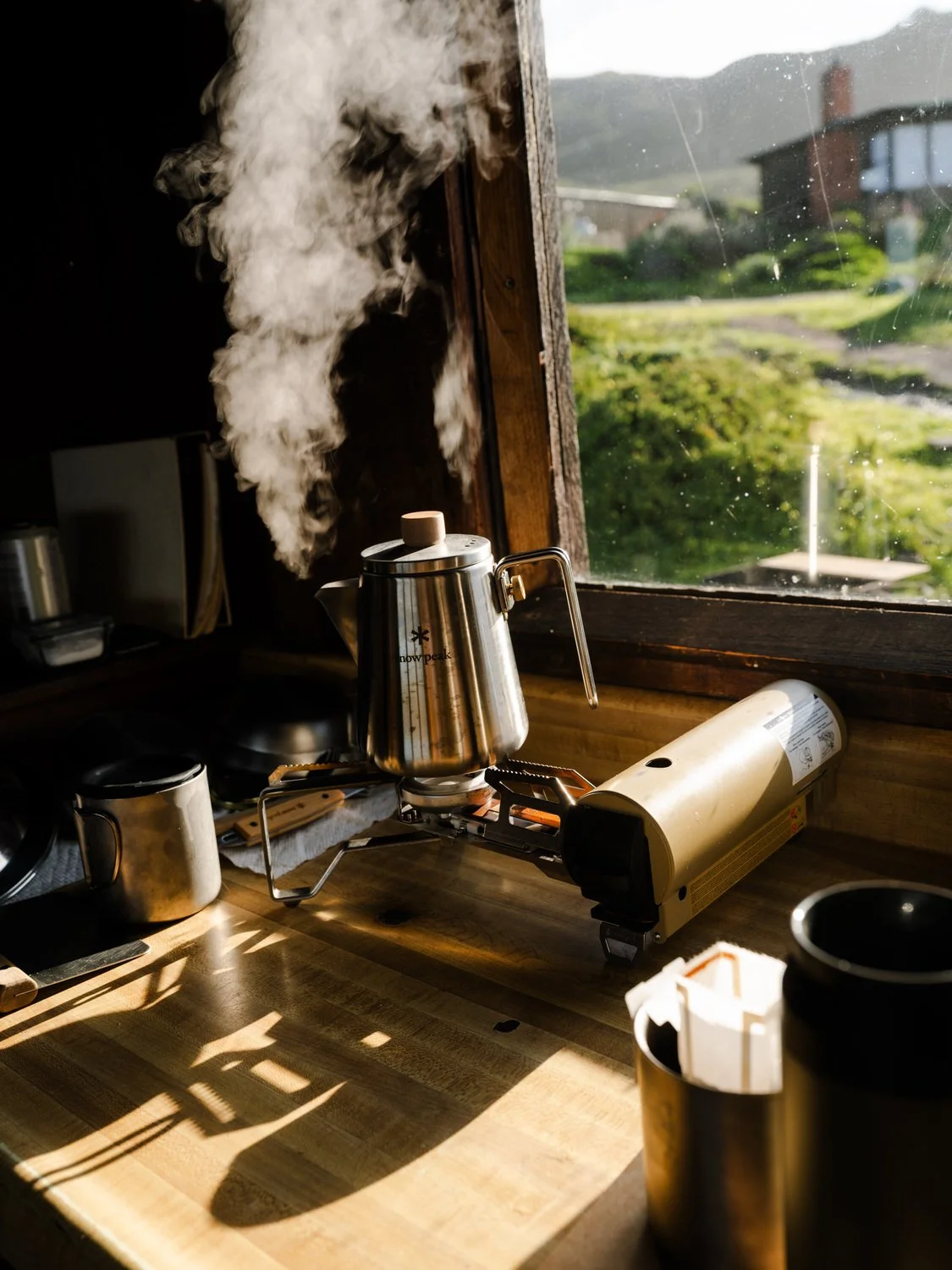 A stainless steel teapot on a small portable stove on a wooden kitchen counter next to a toaster oven, with steam rising from the teapot, and sunlight streaming through a window showing a grassy yard and houses outside.