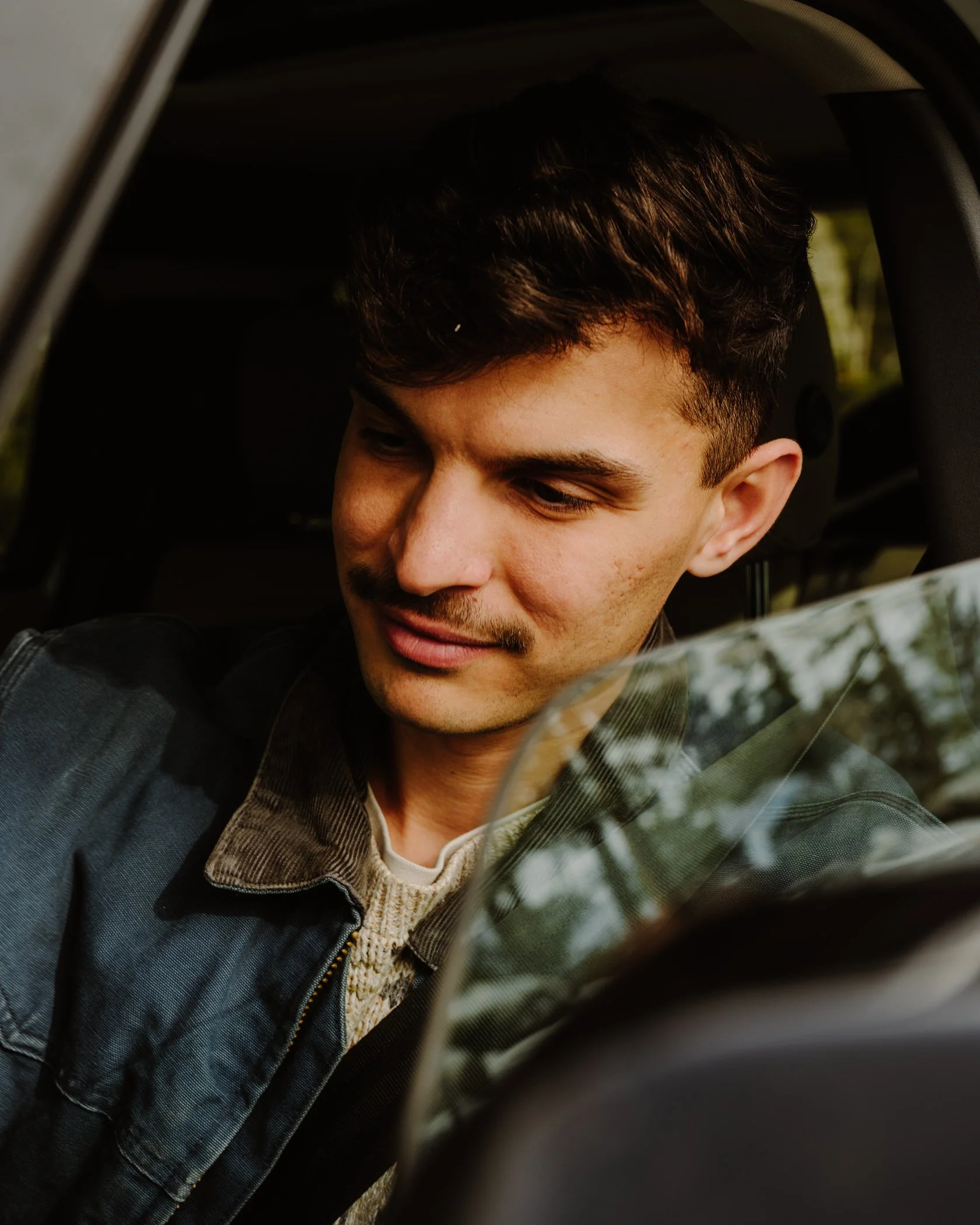 Close-up of a young man with short, dark hair, smiling, wearing a denim jacket over a beige knit sweater, sitting in a vehicle.