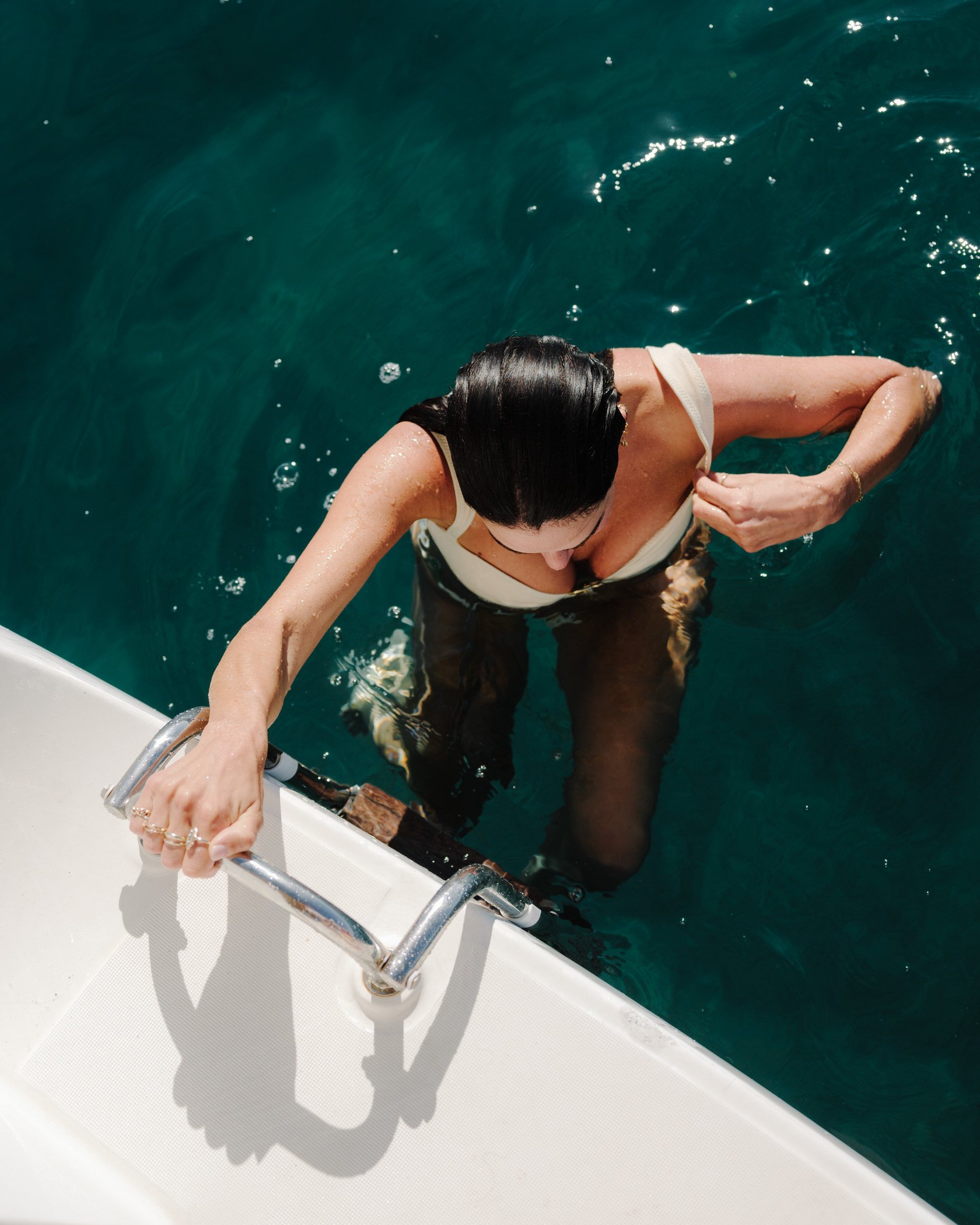 A person with long dark hair climbing out of the water using a boat ladder, with their back partially submerged and their right arm grabbing the ladder.