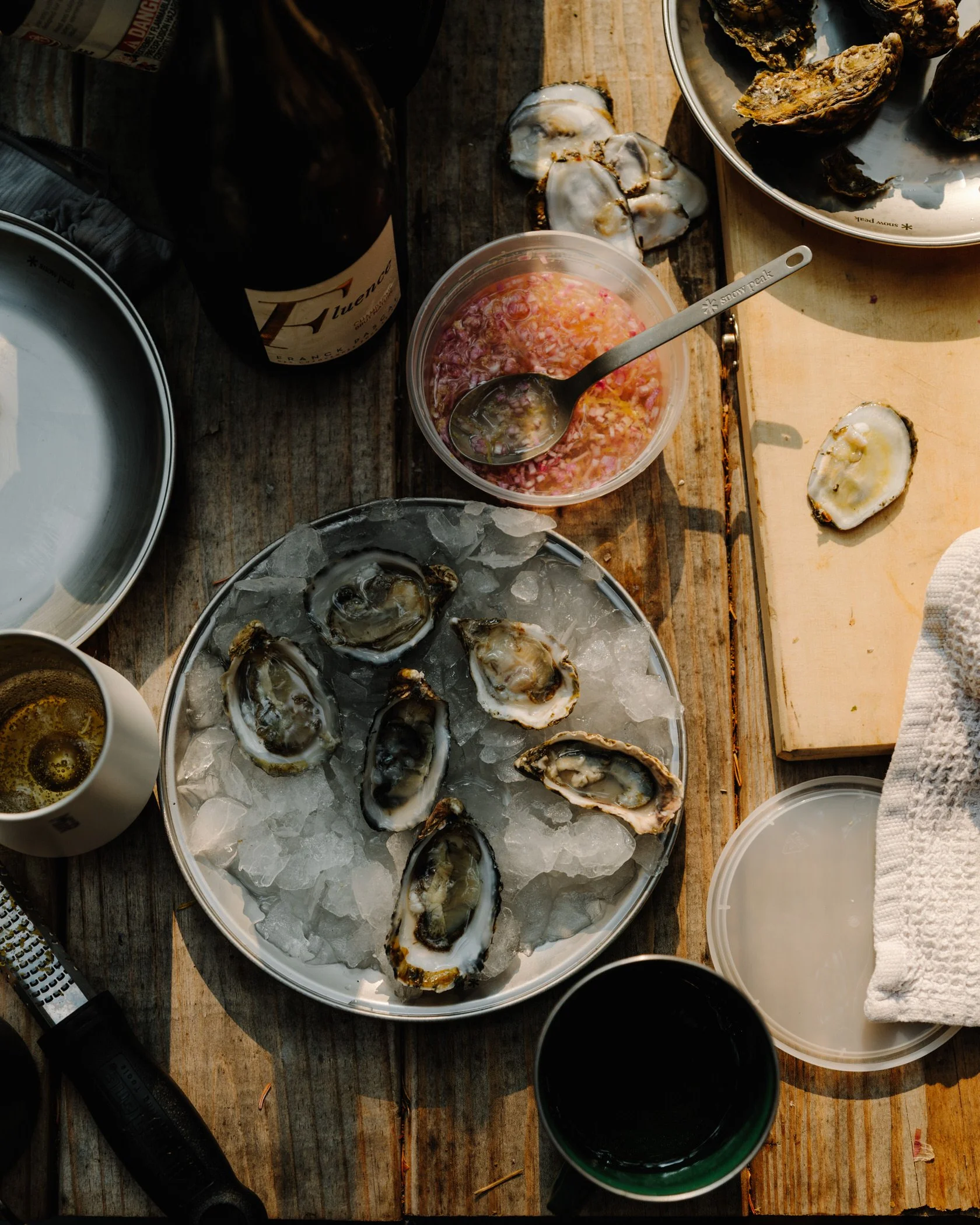 Raw oysters on ice, a bowl of chopped seafood mixture, open oyster shells, a bottle of wine or beer, a black flashlight, a white towel, and containers of sauce on a rustic wooden table.