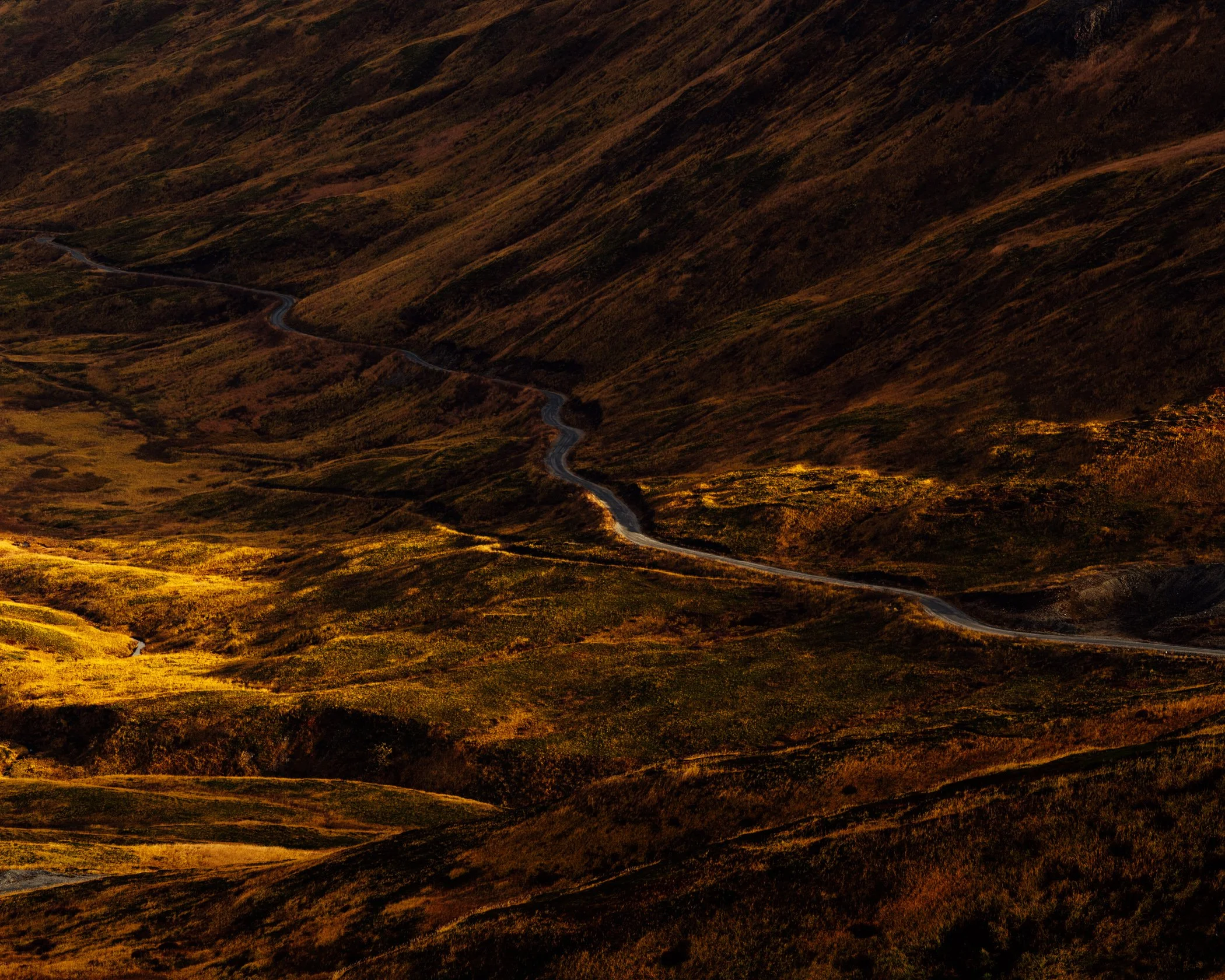 A winding road runs through a lush green valley surrounded by steep hills with trees and vegetation, in what appears to be a remote mountainous area during late afternoon or early evening.
