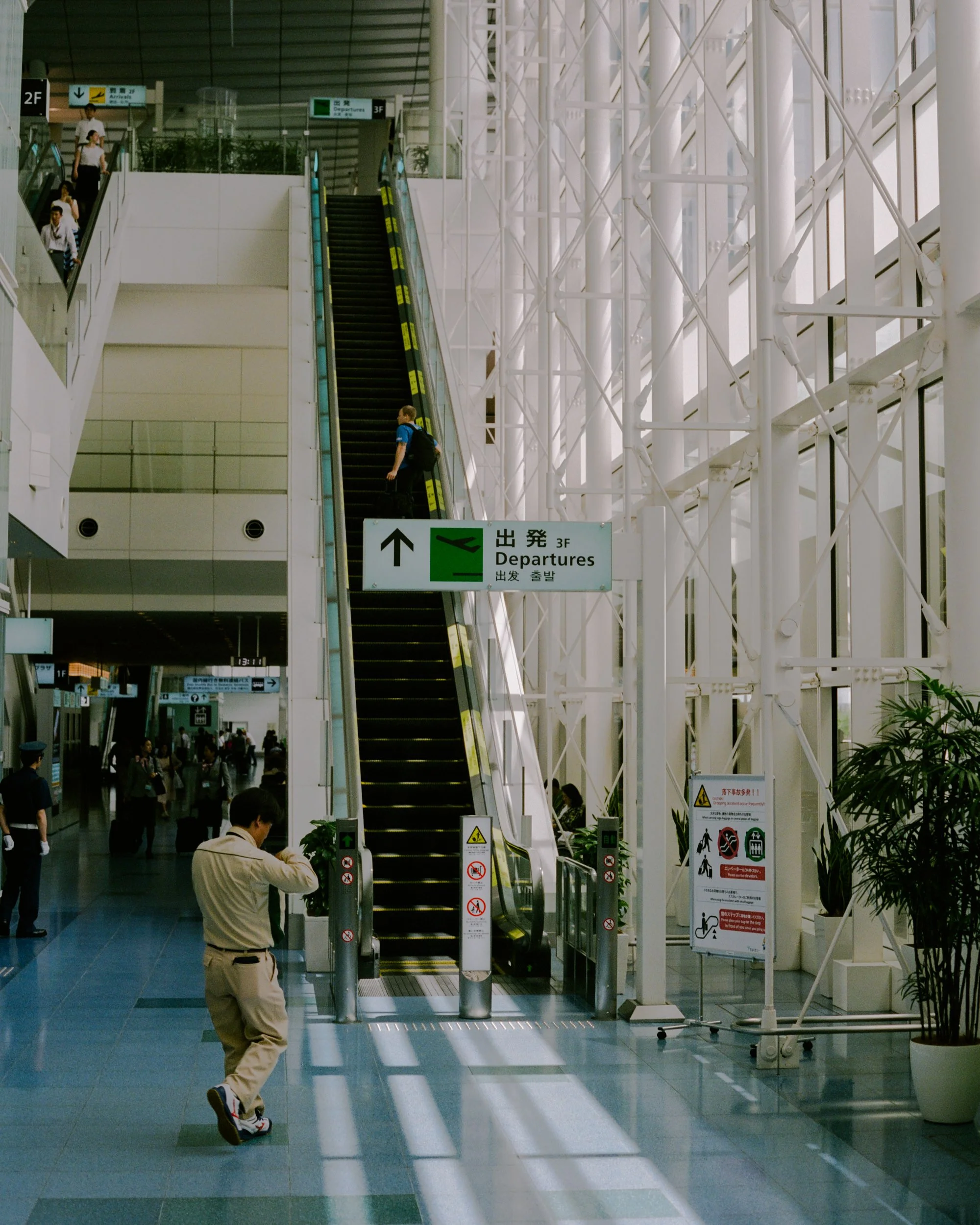 An airport interior with an escalator in the center, people waiting and walking around, signs for departures on the second and third floors, and a man in a white jacket standing near the escalator.