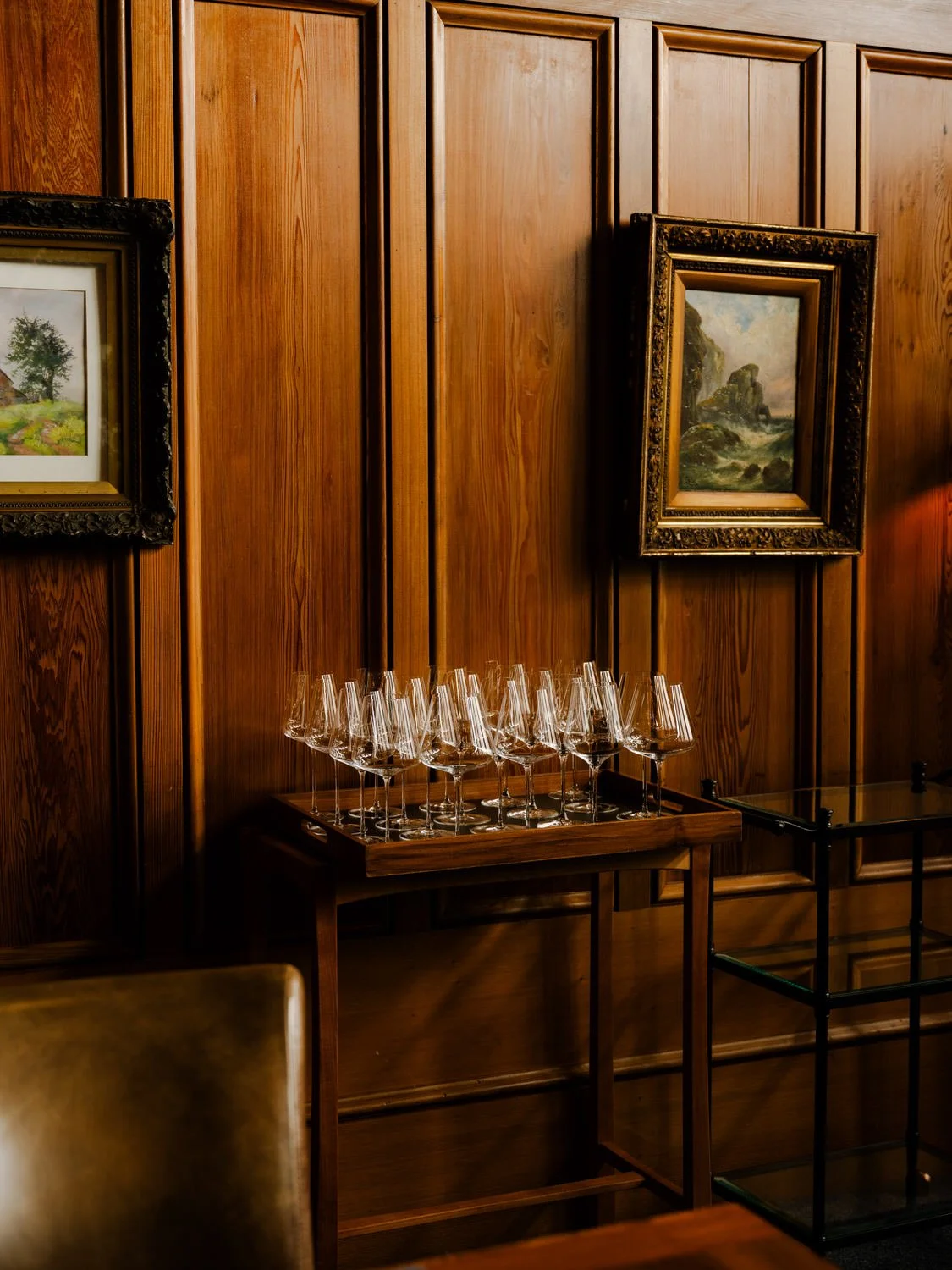 A wooden wall with two framed landscape paintings, a wooden tray with empty wine glasses on a stand, and a small black glass table.