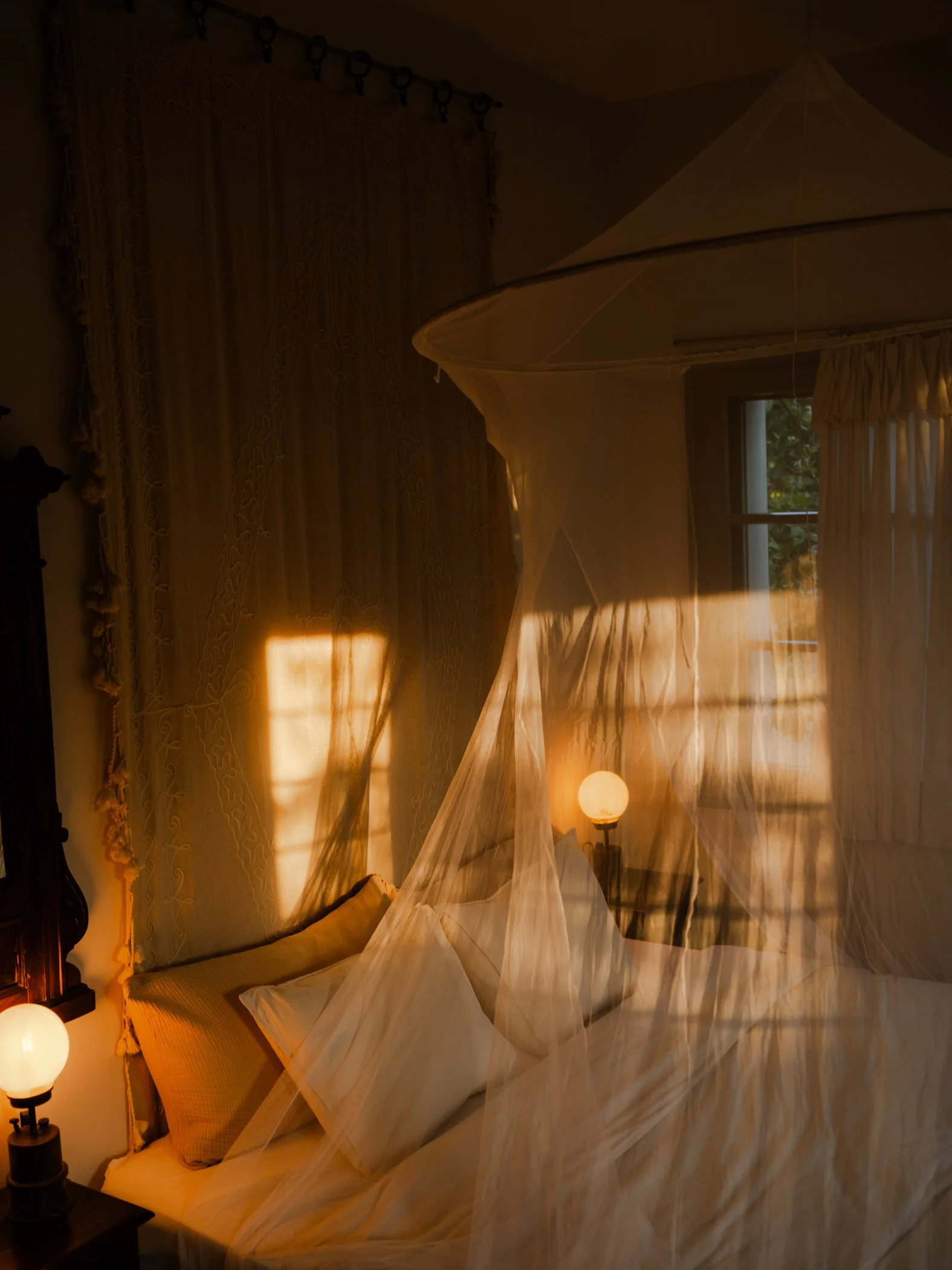 A bedroom with a large window and a bed with yellow pillows, illuminated by warm lighting from lamps, with a sheer curtain and a decorative drapery.