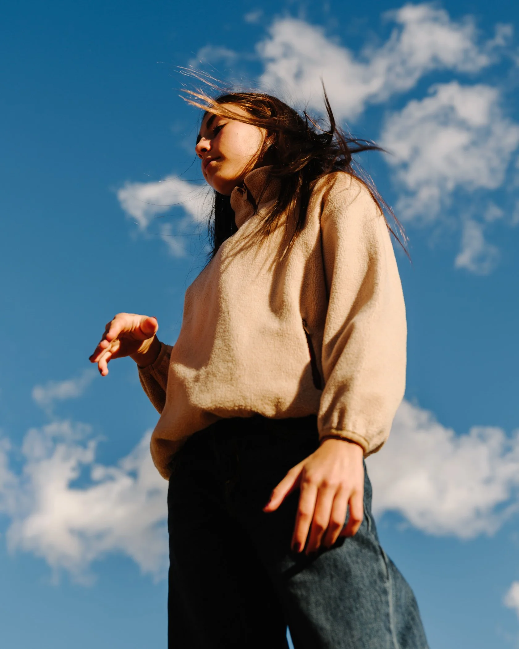 Close-up of a woman standing outdoors against a blue sky with clouds, wearing a beige fleece jacket and jeans.