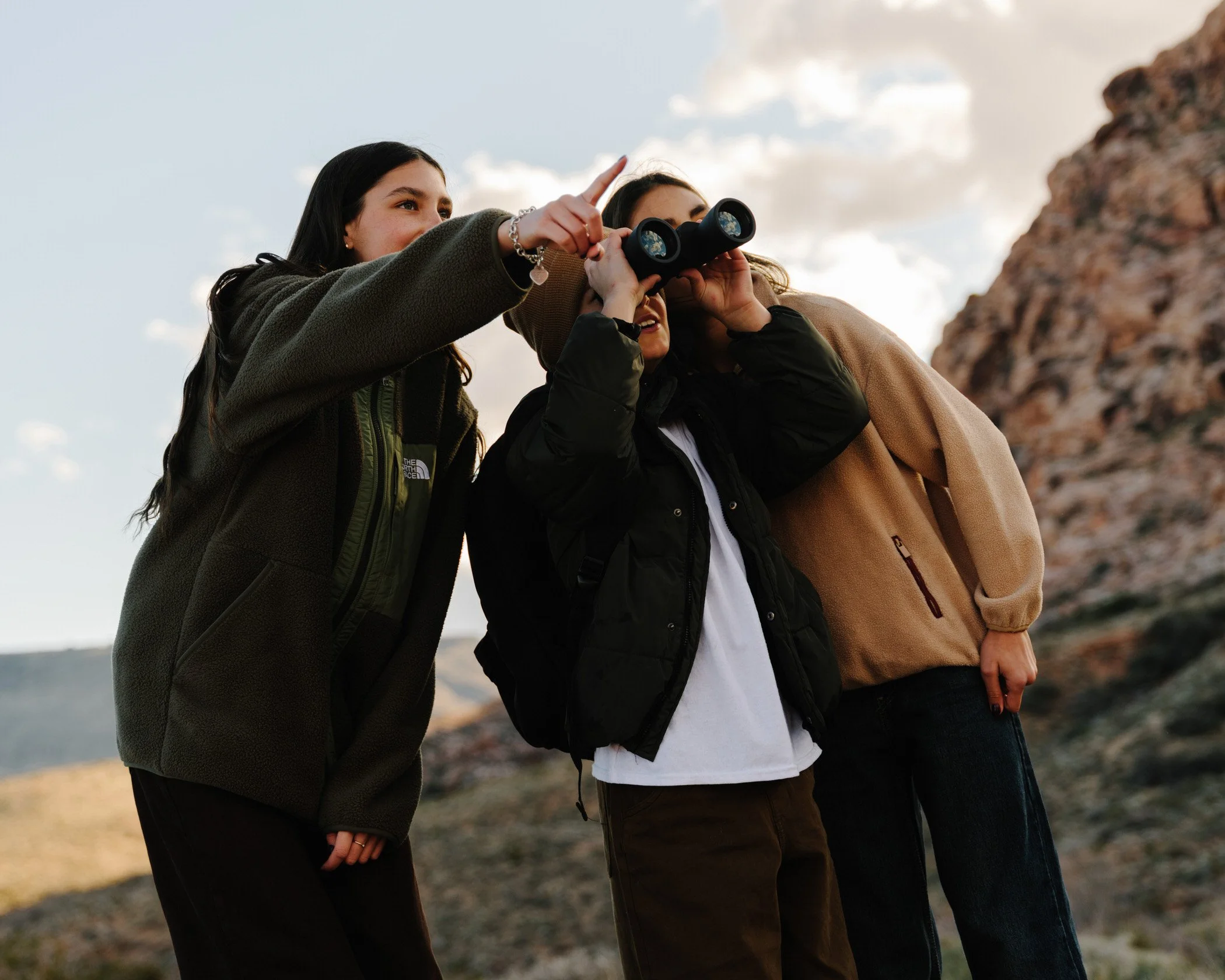 Three women standing outdoors, with one holding binoculars and pointing at something, during sunset or sunrise, with a mountain and cloudy sky in the background.
