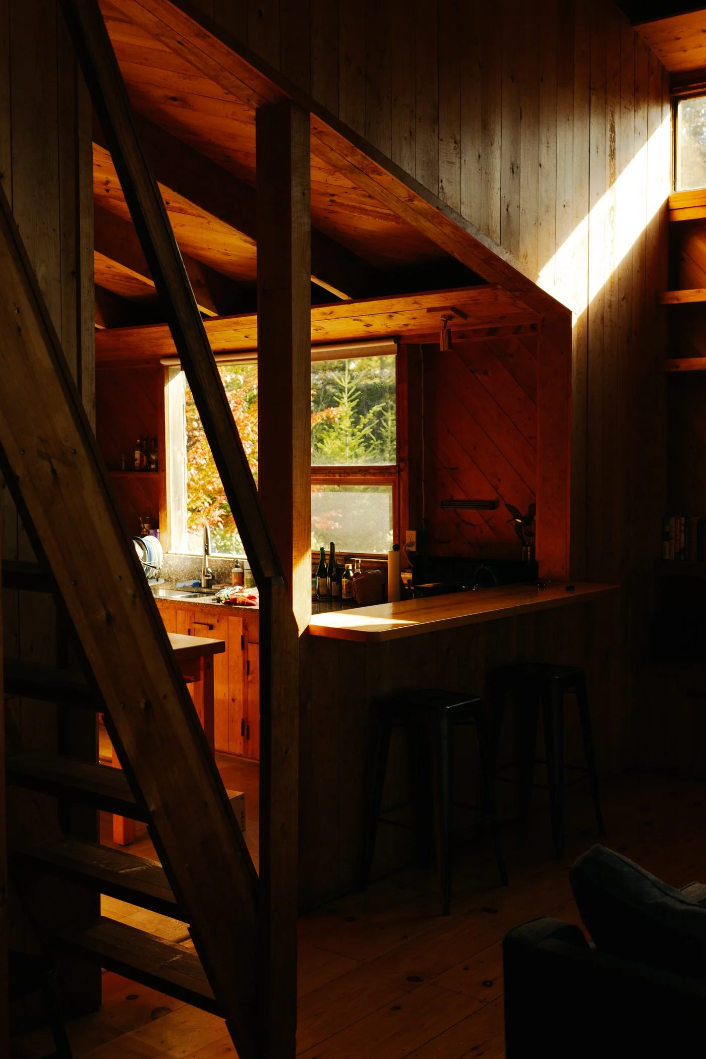Interior of a rustic wooden cabin kitchen, with sunlight streaming through a window, wooden beams, and a countertop with bottles and kitchen items.