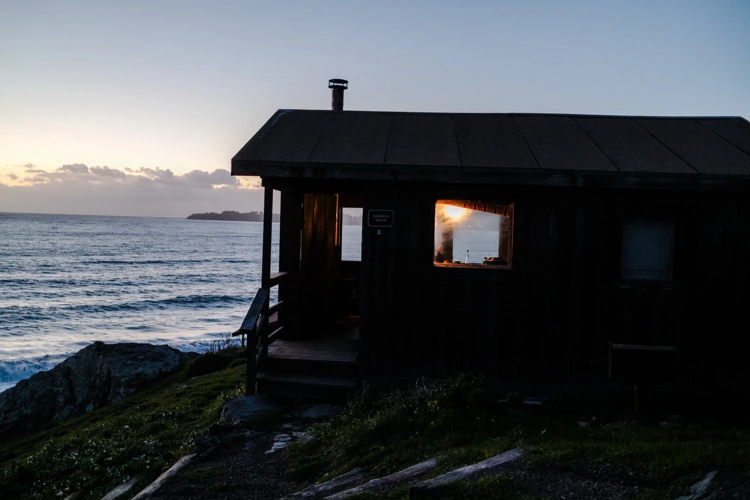 A small wooden house on a cliff overlooking the ocean at sunset, with sunlight visible through a window.