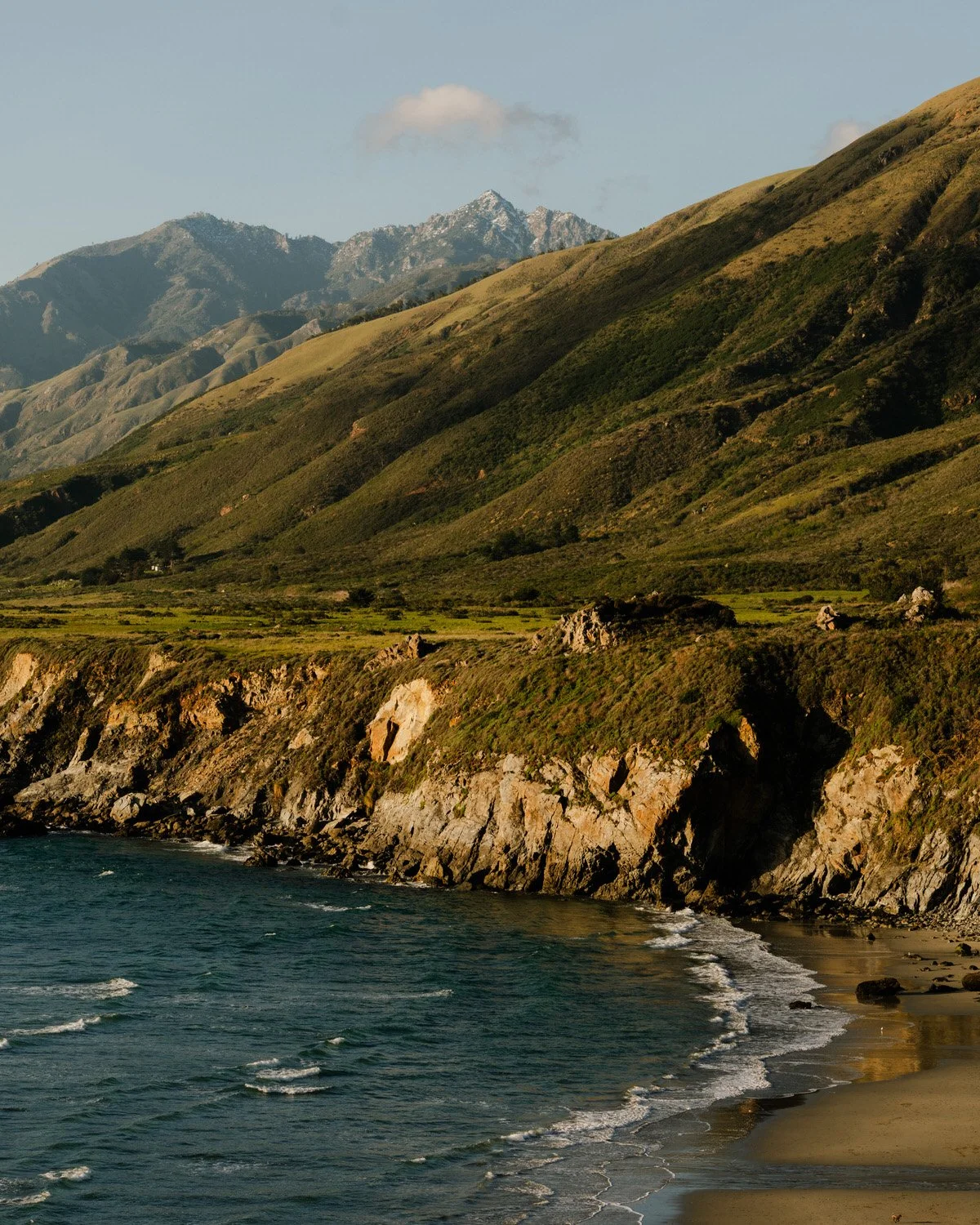Coastal landscape with rocky cliffs, green hills, and mountains in the background, ocean in the foreground, and a cloudy sky.