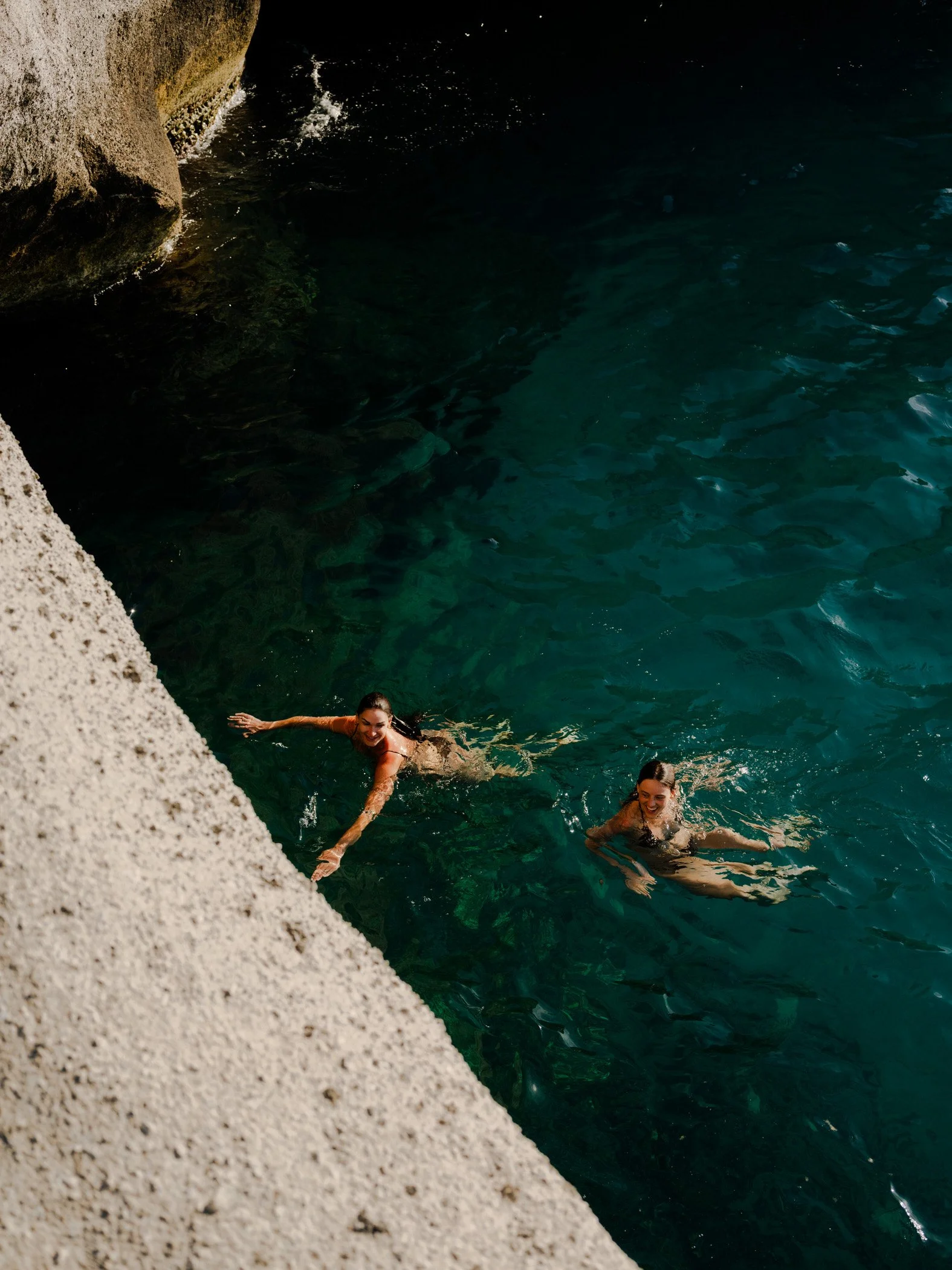 Two women swimming in the water near a rocky shoreline at sunset.