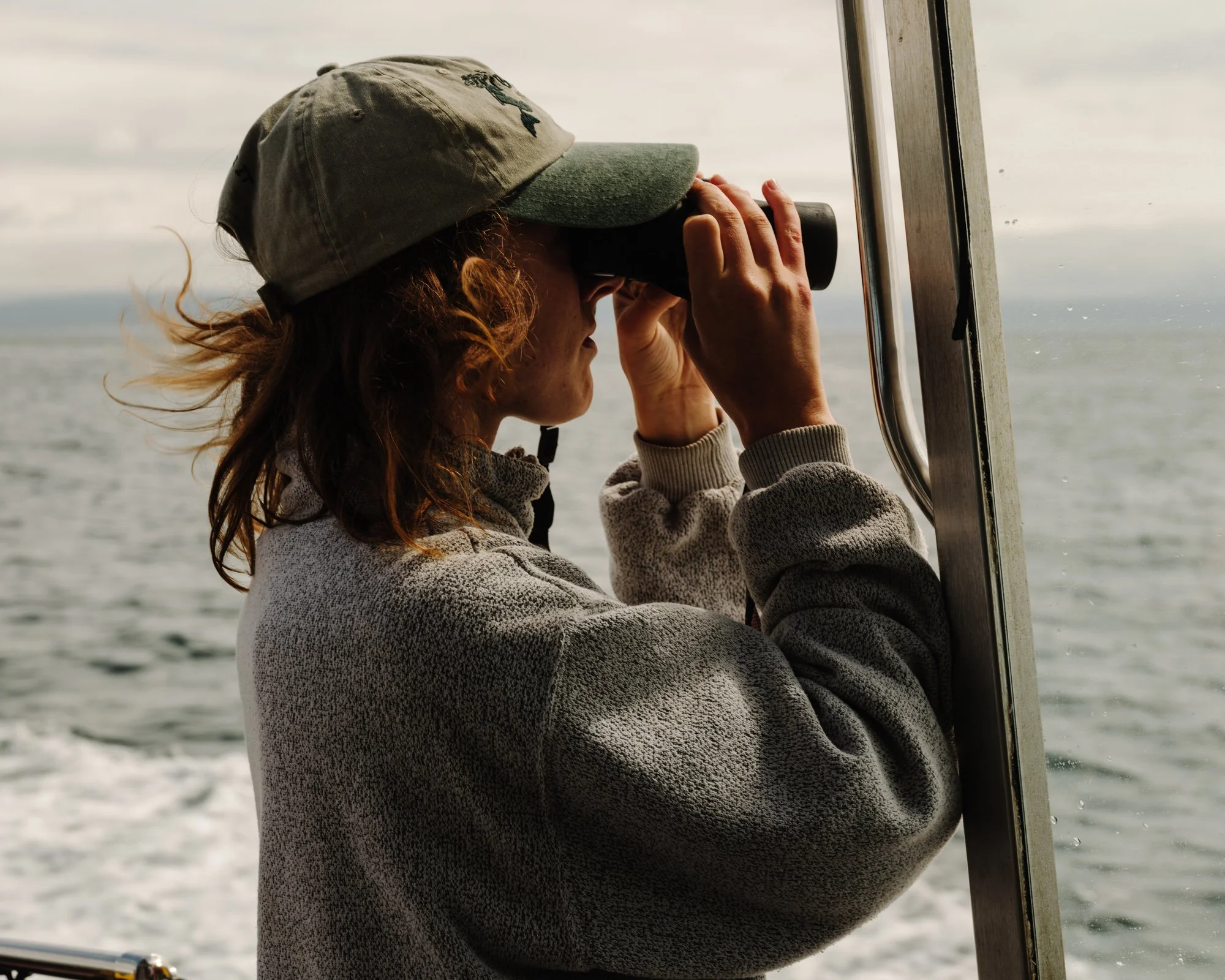 A person with curly hair wearing a gray hat and sweatshirt looks through binoculars at sea from a boat.