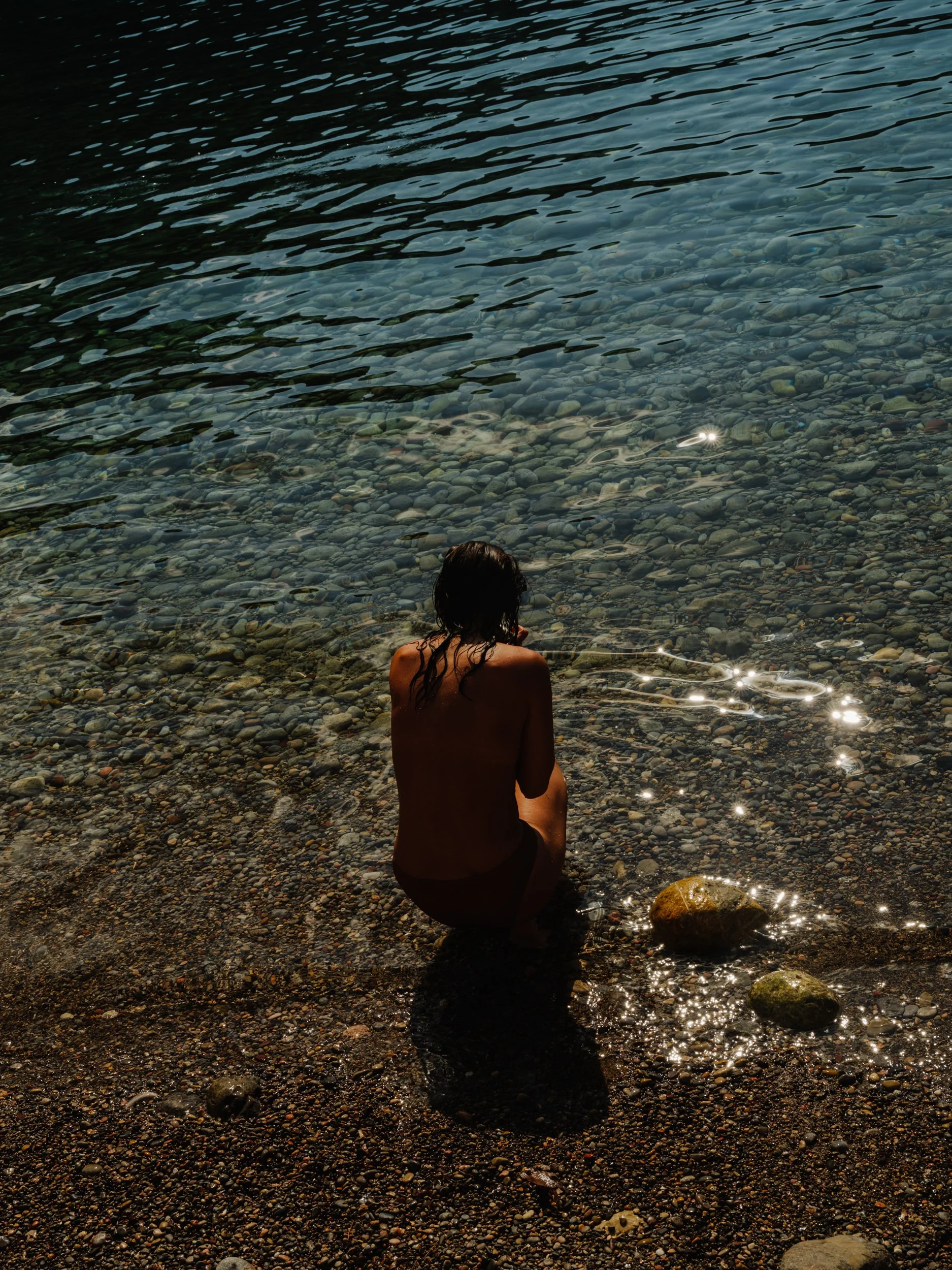 A person with long dark hair sitting on a rocky beach facing the water, with back to the camera, surrounded by pebbles and rocks, illuminated by sunlight reflected on the water.