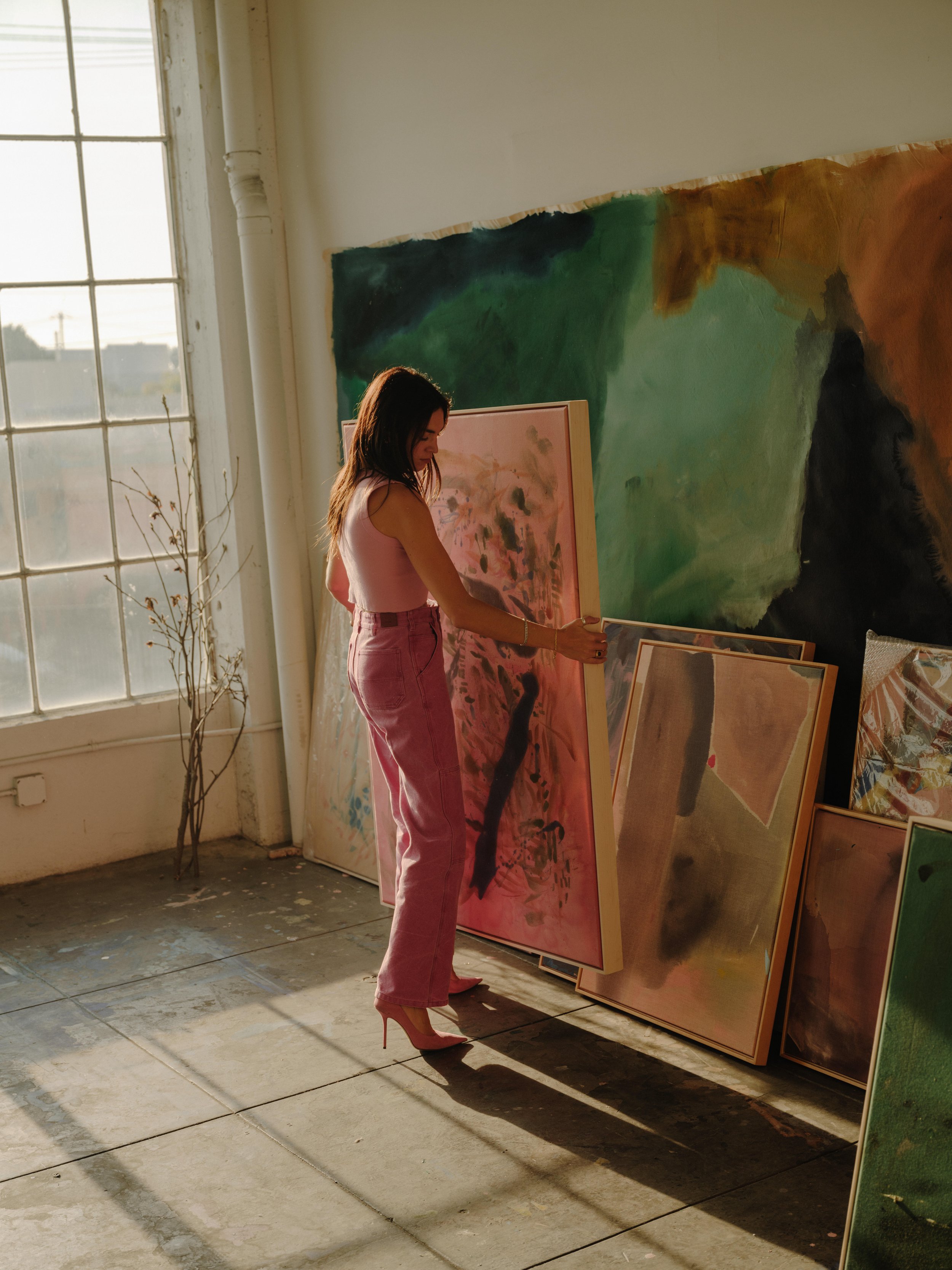 A woman with long dark hair wearing pink high heels, a sleeveless pink top, and pink pants, standing in an art gallery, examining paintings leaning against the wall, with sunlight coming through a large window to her left.
