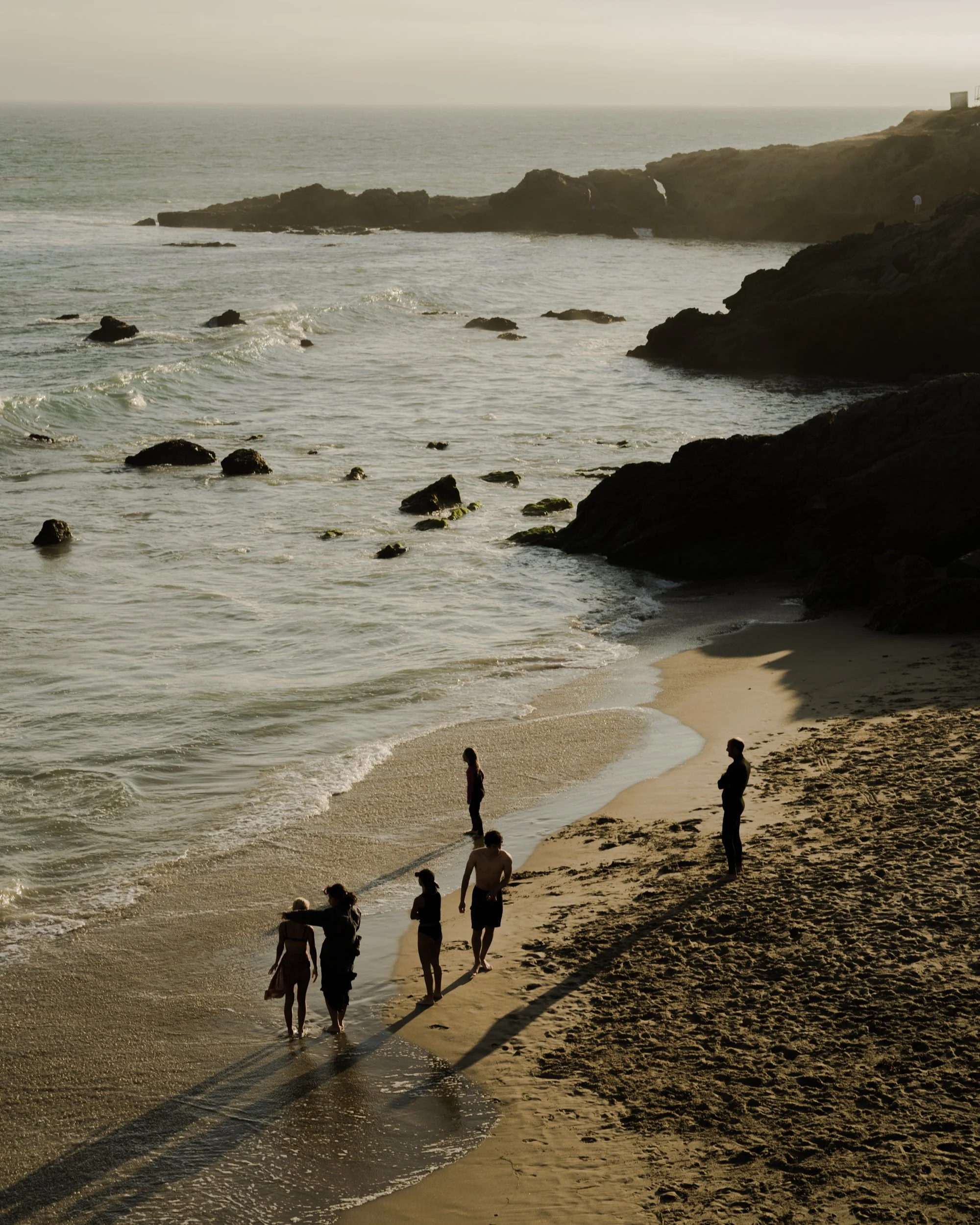 People walking along a sandy beach at sunset, with rocky cliffs in the background and gentle waves in the foreground.