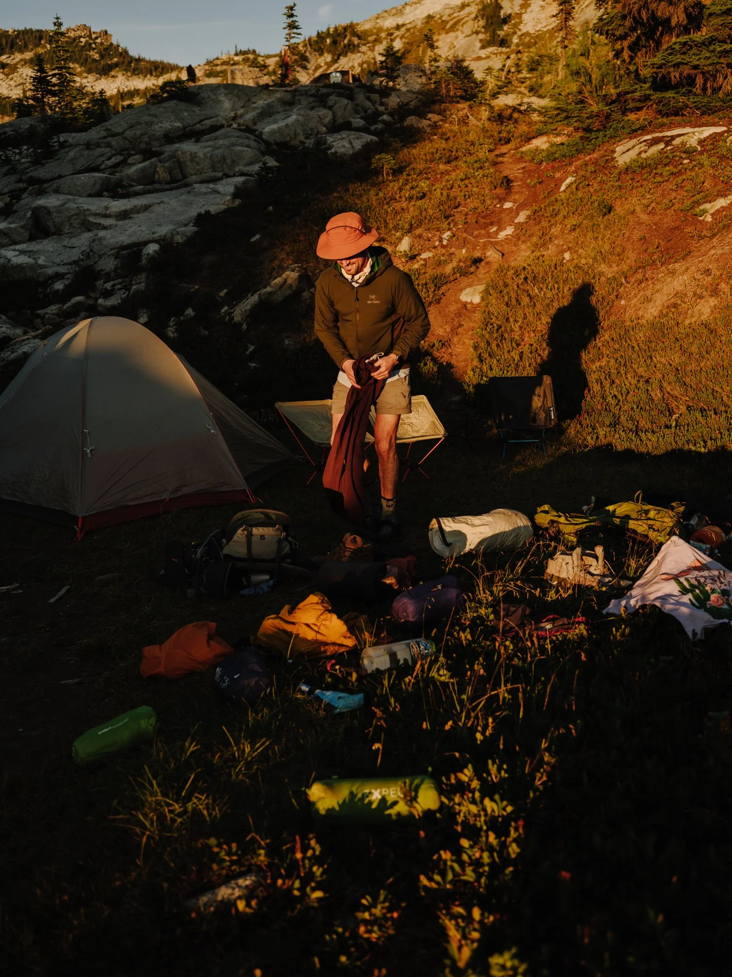 A person standing near a tent outdoors at sunrise or sunset, with camping gear and equipment spread around them in a mountainous, forested area.