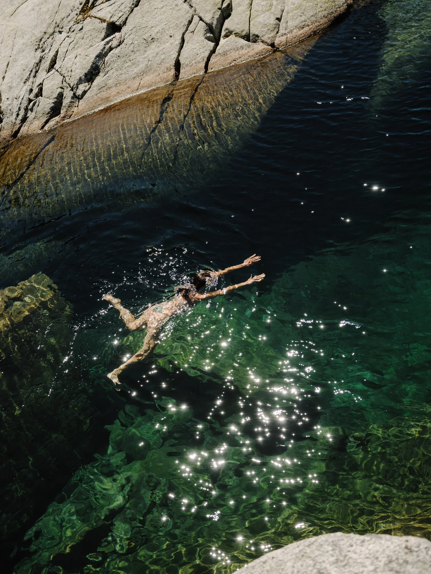 A person swimming in a clear body of water next to rocks, with sunlight reflecting on the water's surface.