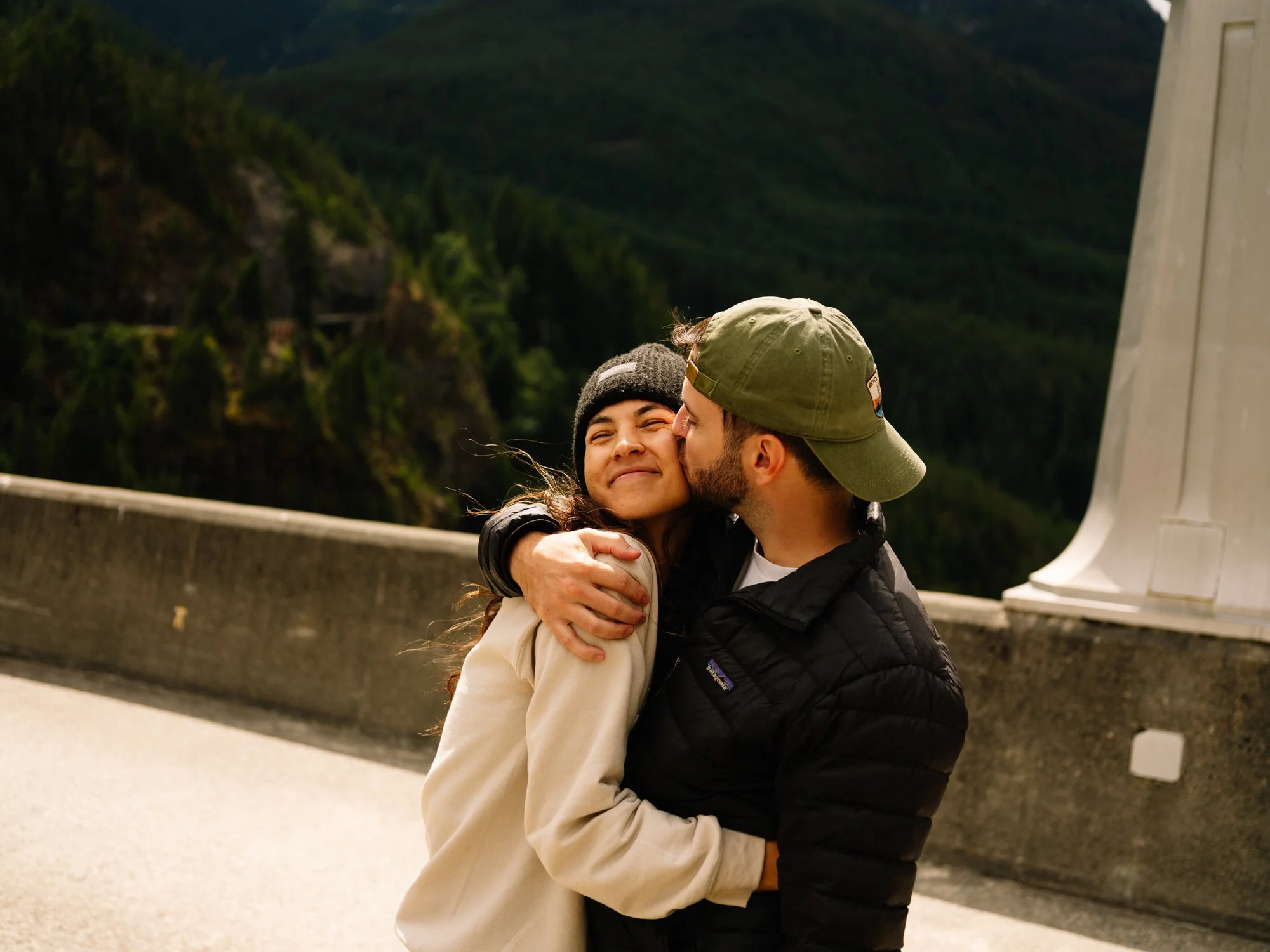 A young woman with curly hair and a beanie being kissed on the cheek by a man wearing a baseball cap, both smiling with a mountain landscape and a bridge in the background.
