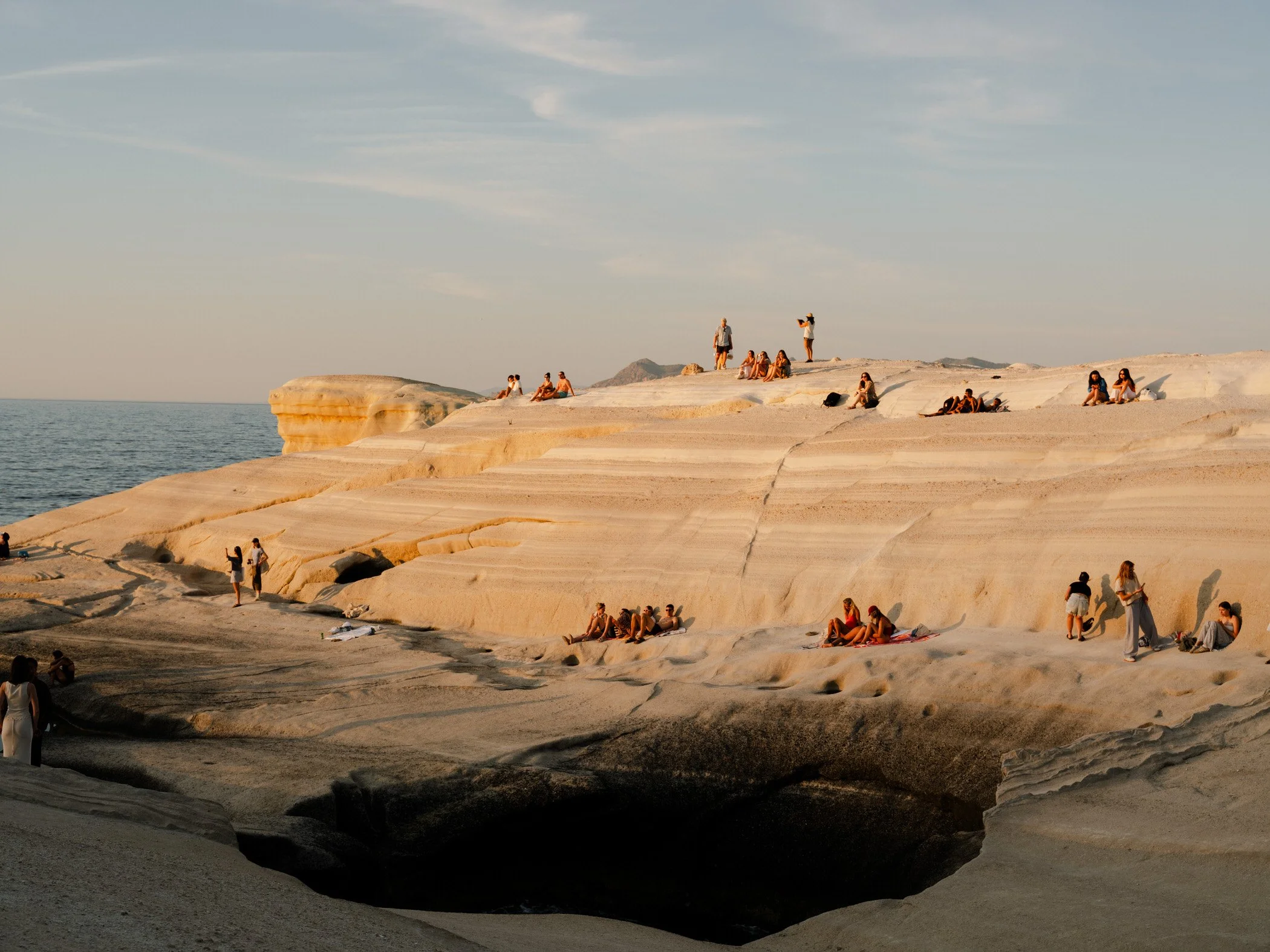 People sitting and standing on layered white rocky formations near the ocean during sunset.