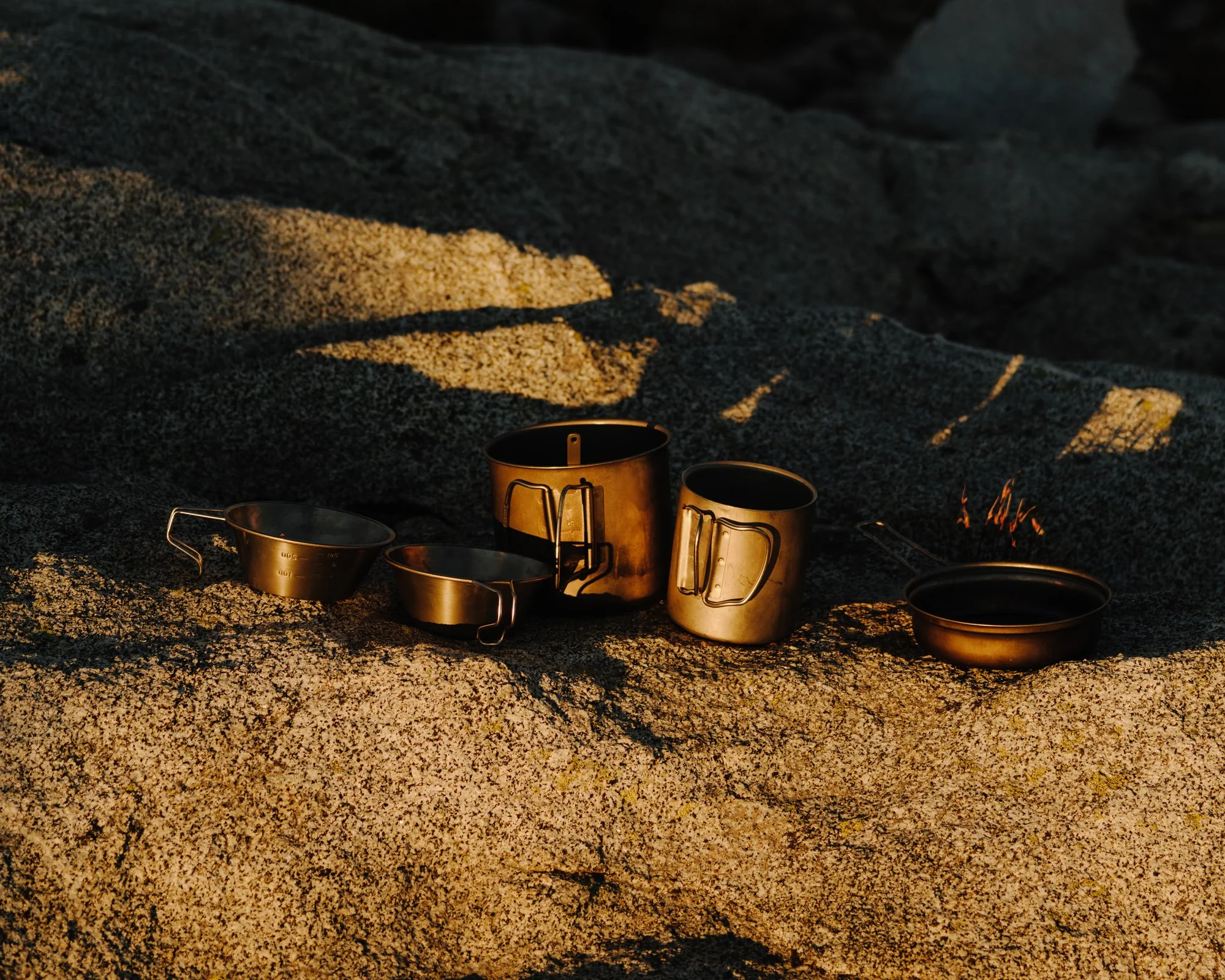 Coffee cups and a small bowl are arranged on a speckled rock surface, with warm sunlight casting shadows.