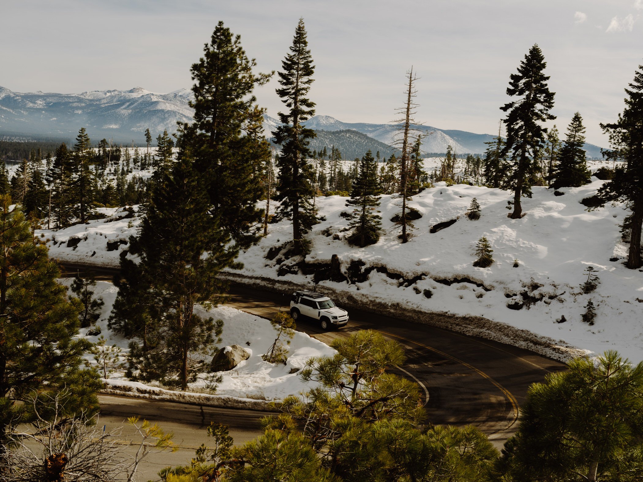 Snow-covered forested mountain landscape with pine trees, snow patches, and a winding road with a white vehicle.