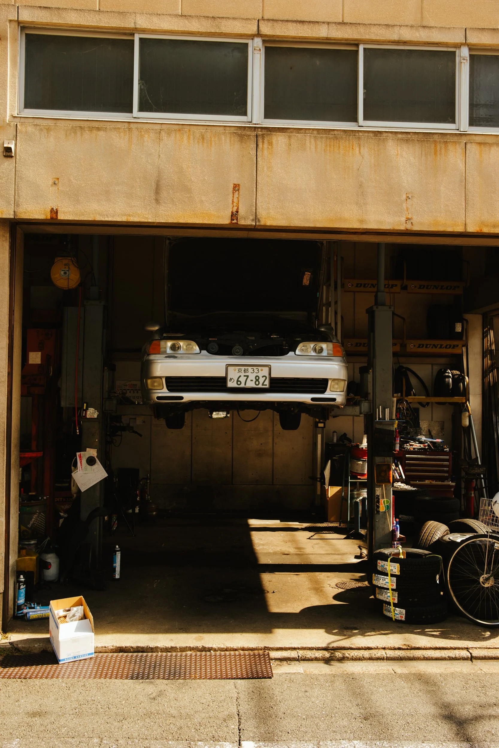 A car partially raised on a hydraulic lift inside an automotive repair shop. The shop has work tools and tires scattered around, with sunlight casting shadows inside.