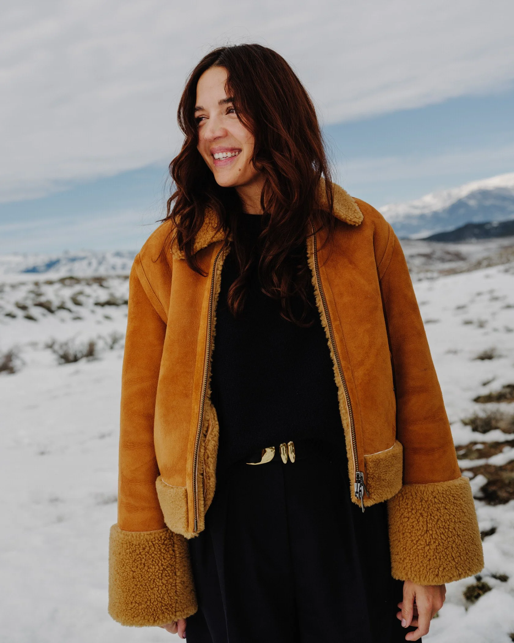A woman with long dark hair wearing a brown shearling coat stands in a snowy landscape with mountains in the background, smiling and looking to the side.