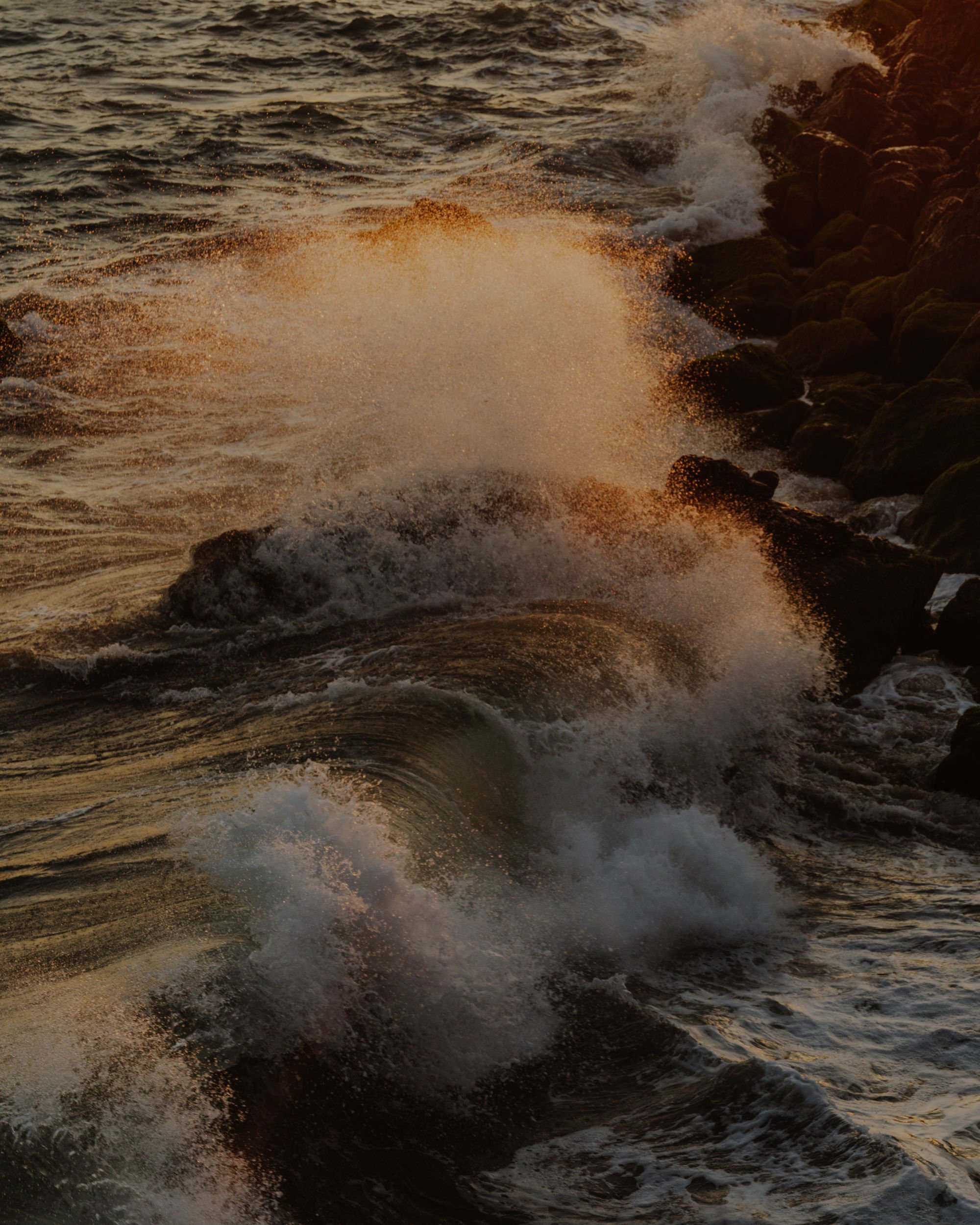 Waves crashing on rocky shoreline during sunset with golden light reflecting off the water.