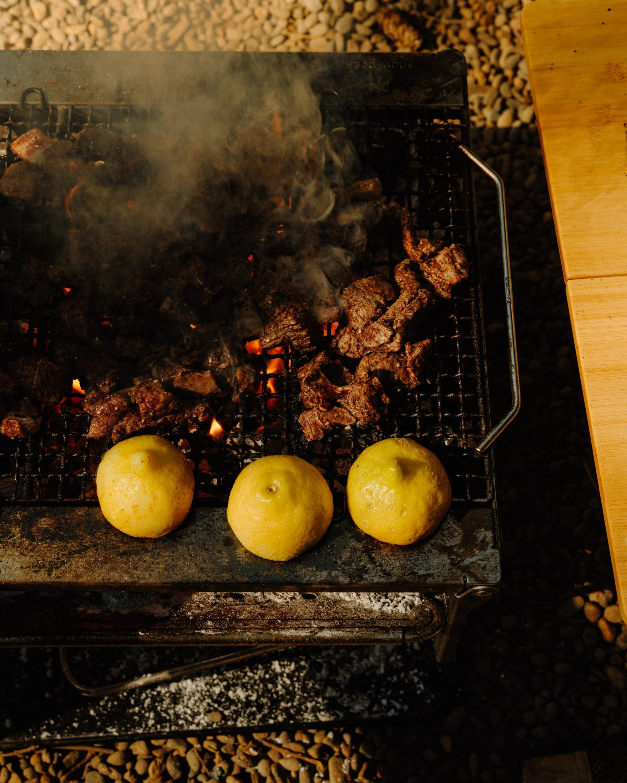 Lemons on a grill with pieces of meat cooking over charcoal, partially covered in smoke.