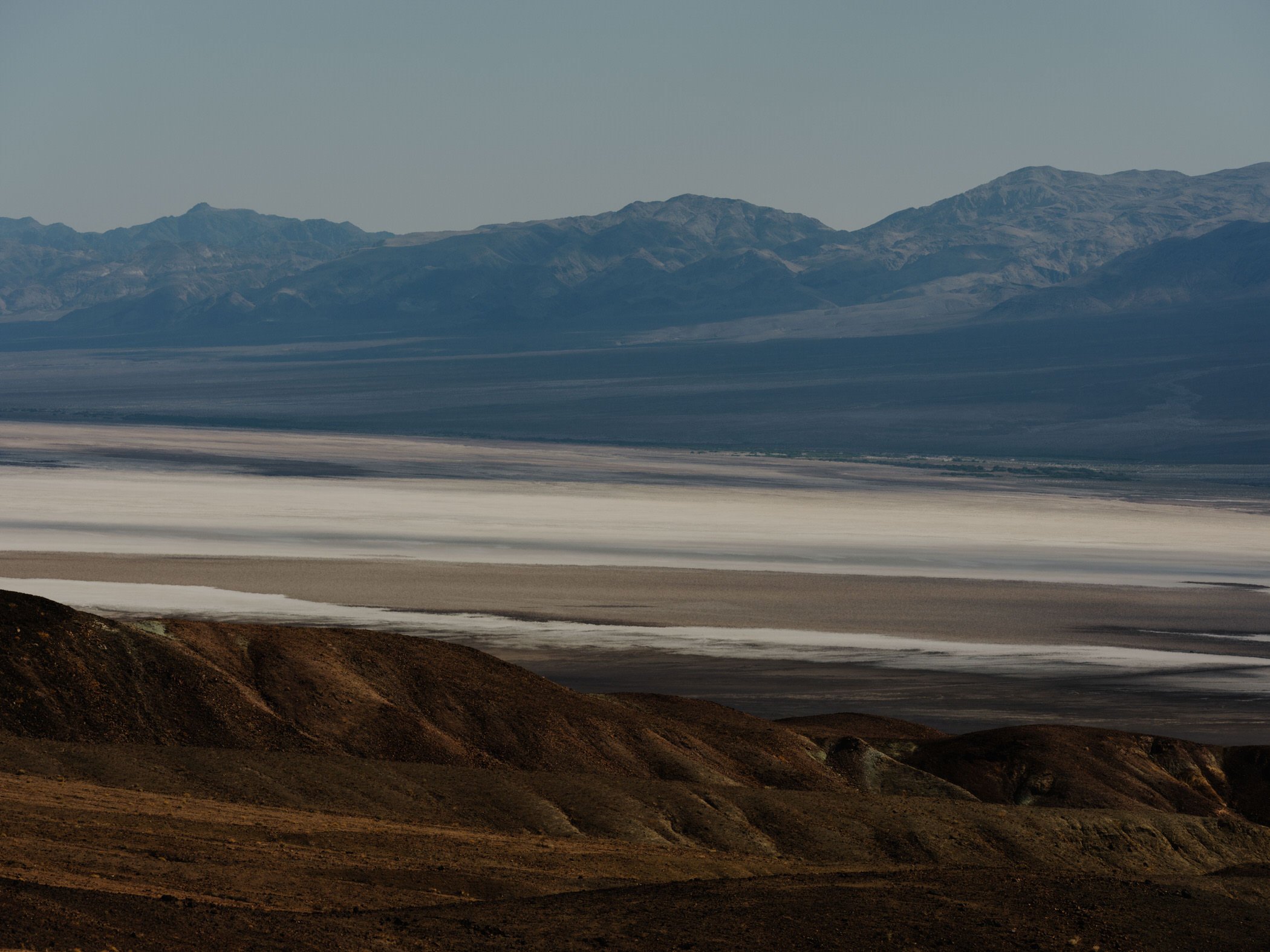 Arid, rugged mountains with brown and dark green slopes and flat plains in the foreground under a clear sky.