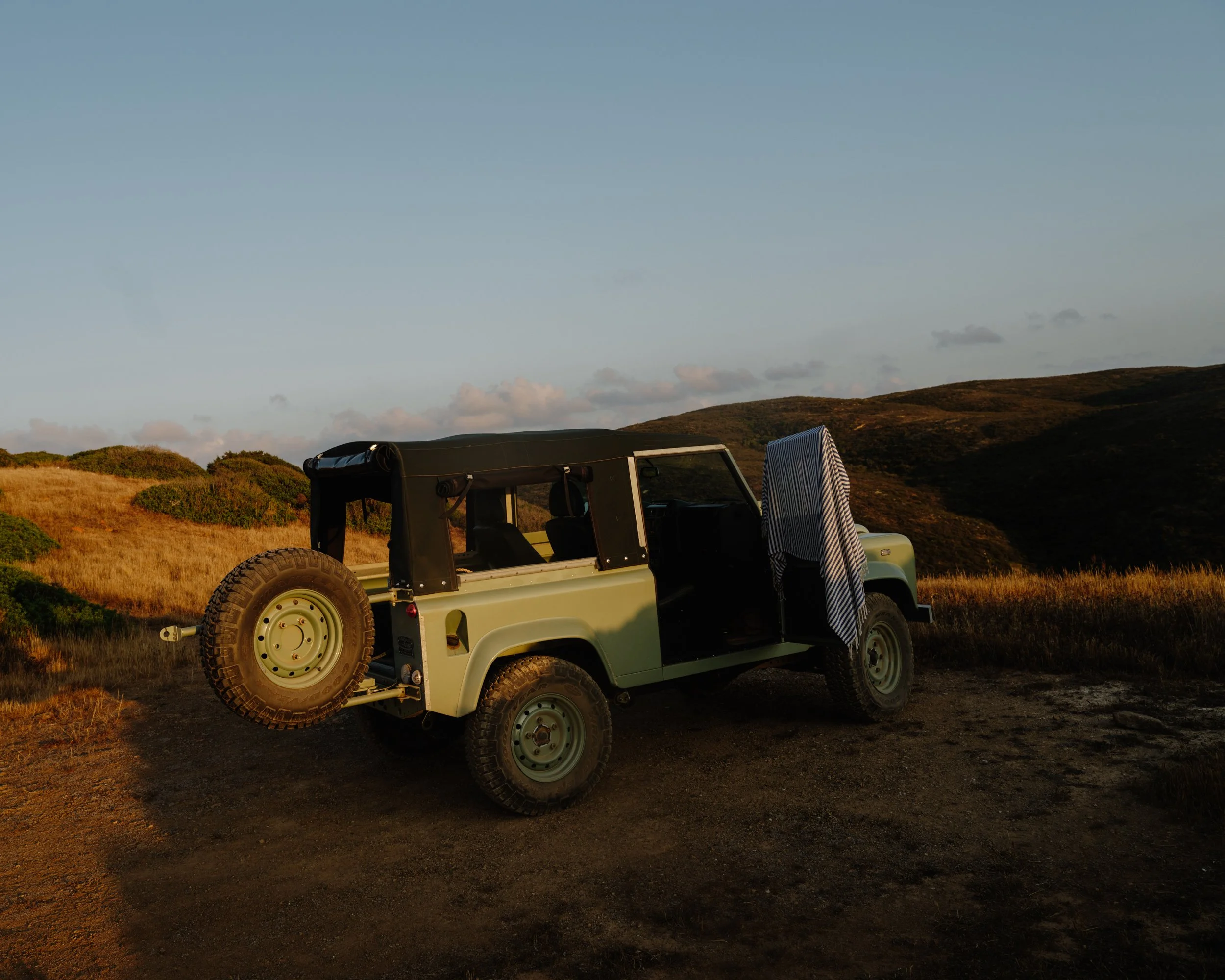 A vintage off-road vehicle parked on a grassy hillside during sunset with hills and a partly cloudy sky in the background.