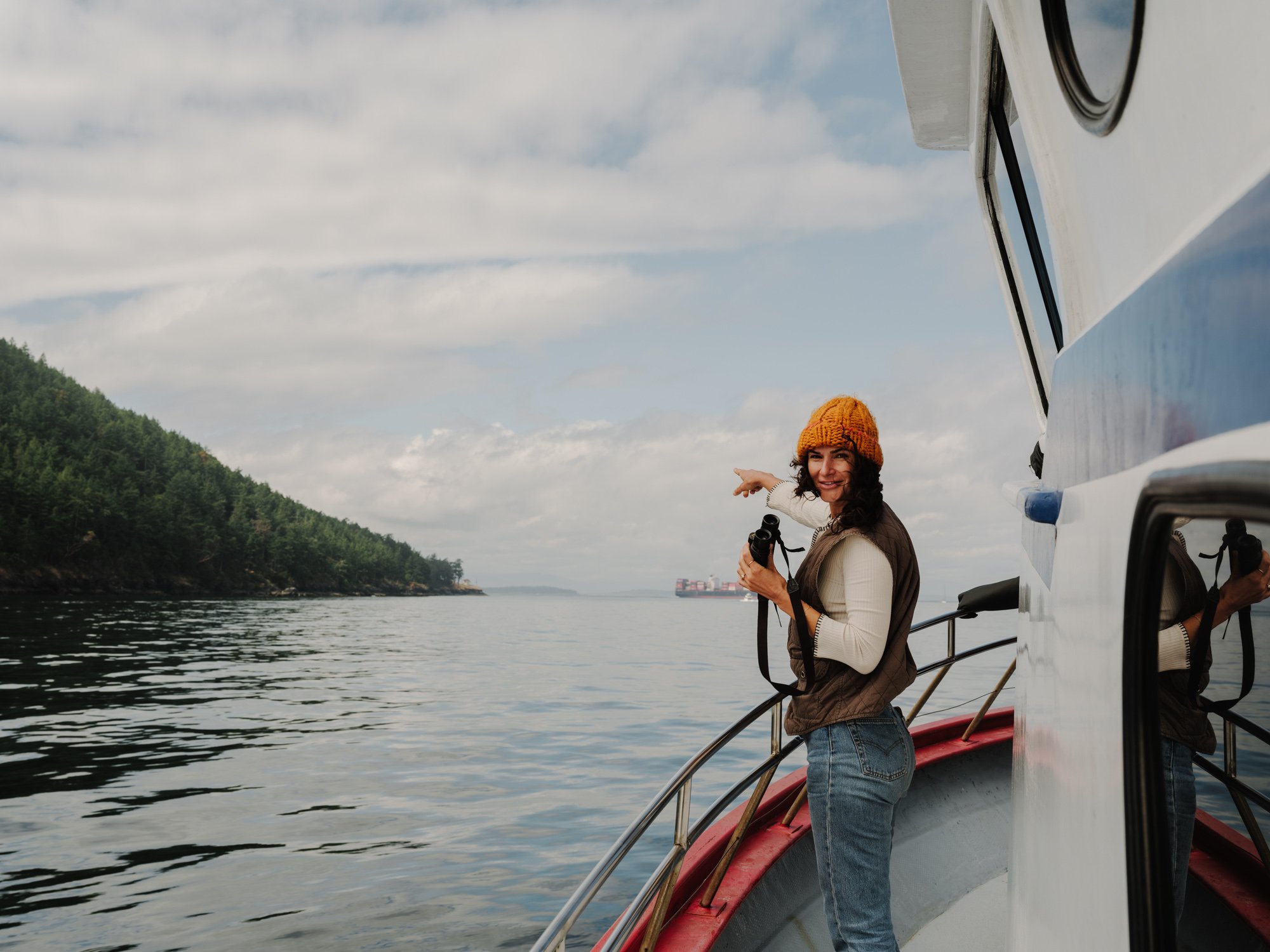 A woman on a boat holding binoculars, wearing a yellow knit hat, white top, and brown vest, pointing at something in the distance over a body of water with green hilly shores.