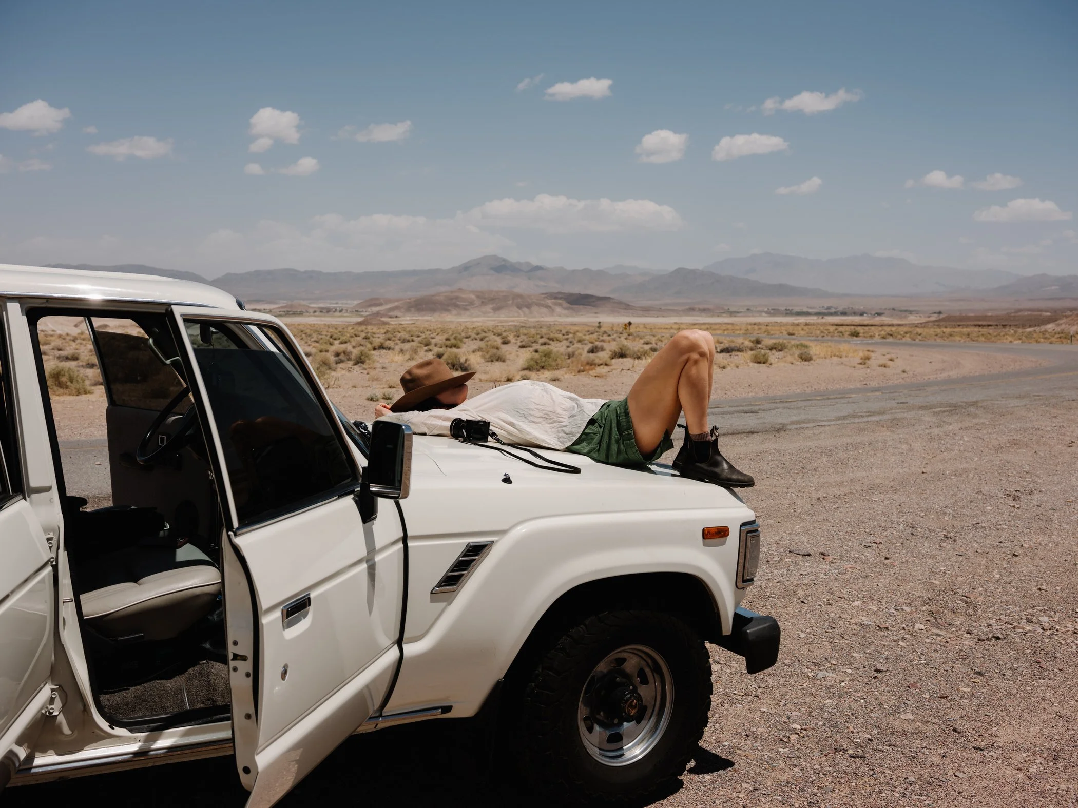 Person lying on the hood of a white vehicle in a desert landscape with mountains in the background, wearing a hat and shorts.