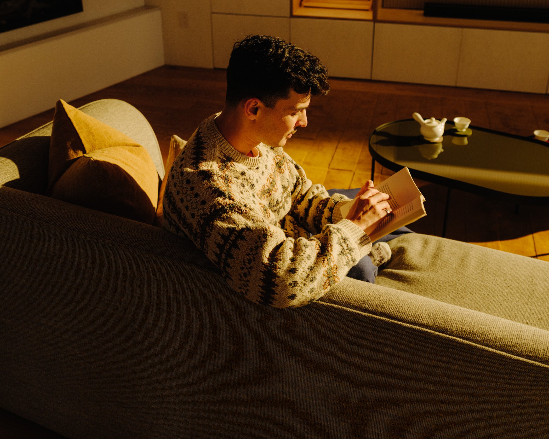 A young man with dark, curly hair sitting on a sofa, reading a book in a warmly lit living room with wood paneling. There are decorative pillows and a black coffee table with a white teapot and cups on it.