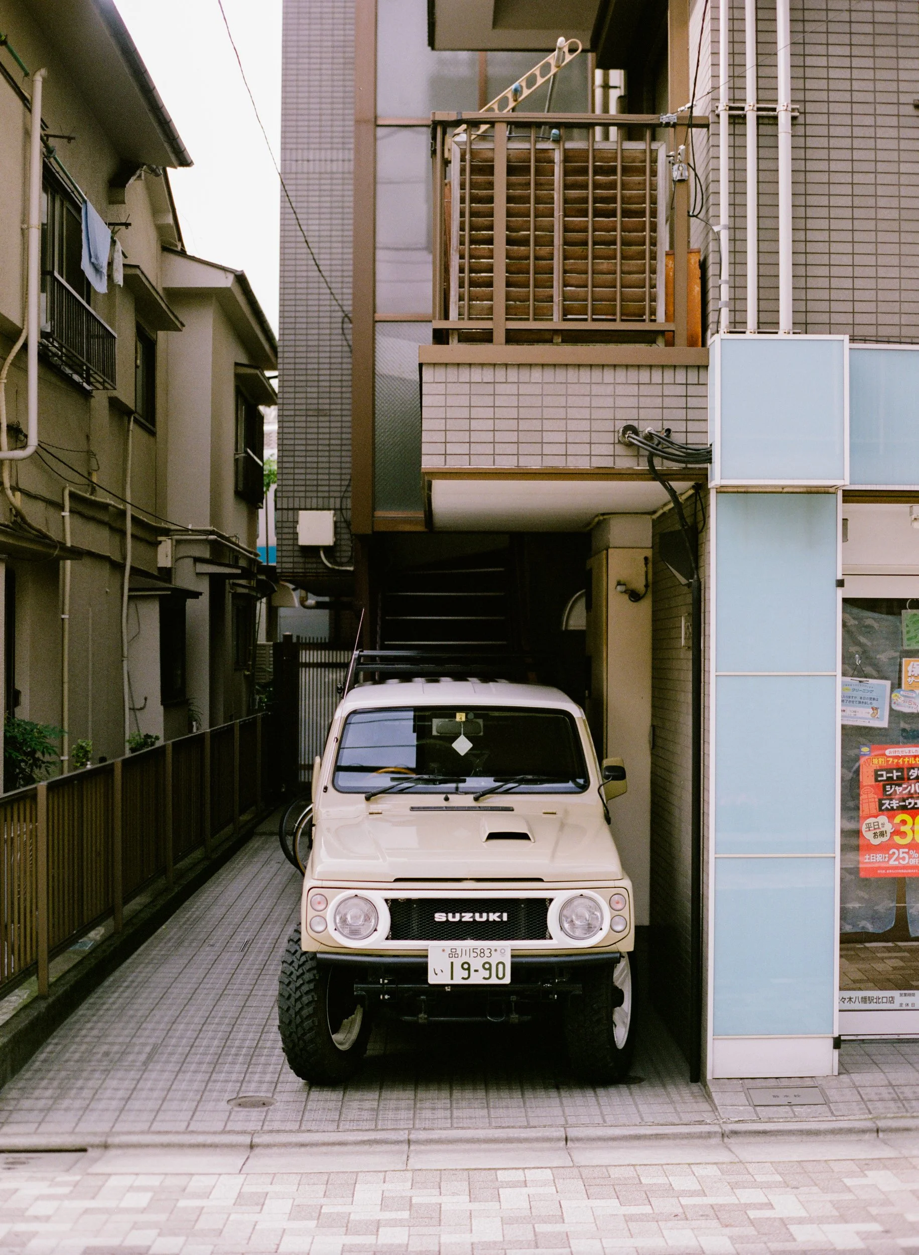 A white Suzuki off-road vehicle parked in a narrow outdoor space between buildings, with a Japanese license plate visible.