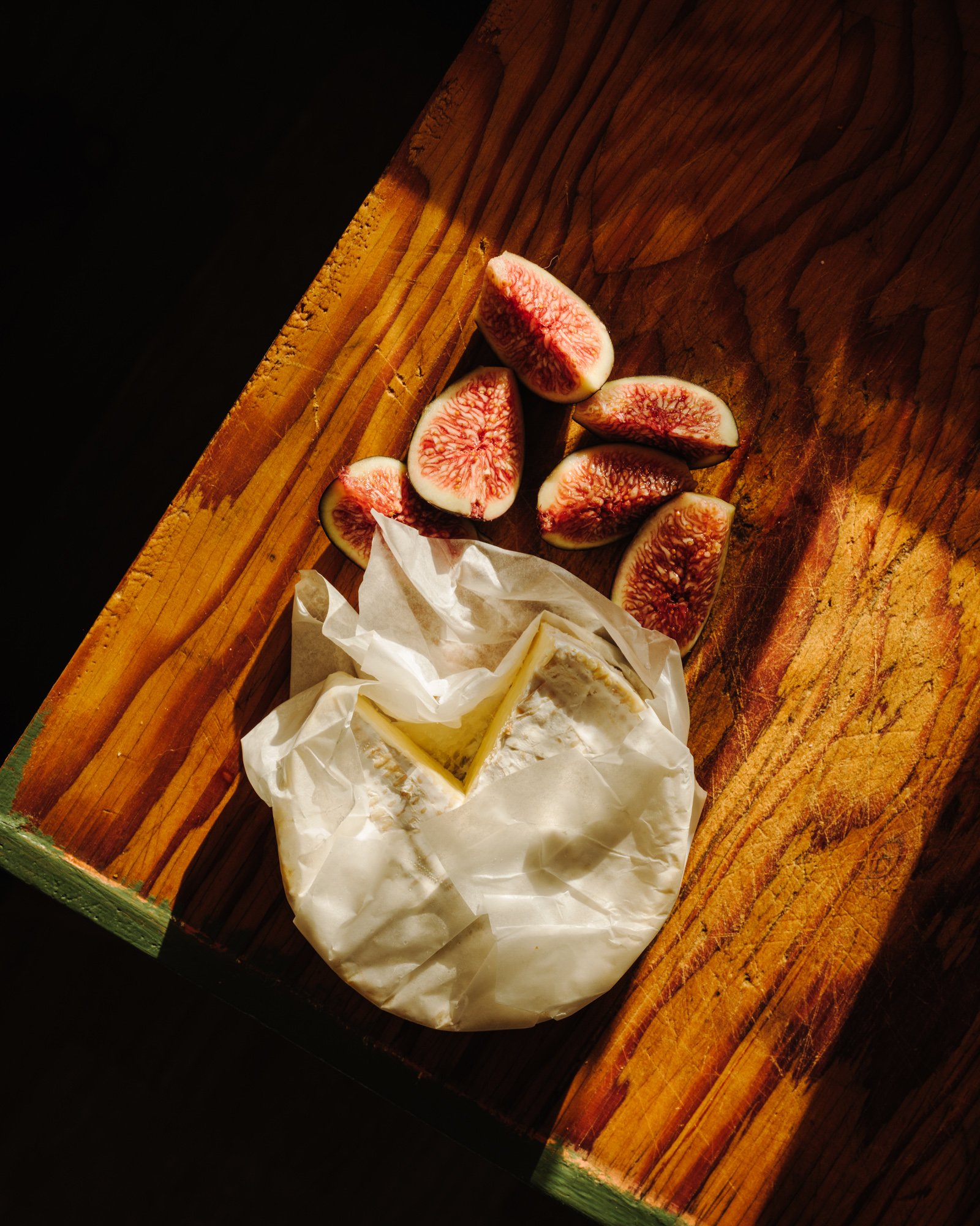 Figs cut in half on a wooden surface, with some cheese wrapped in paper nearby.