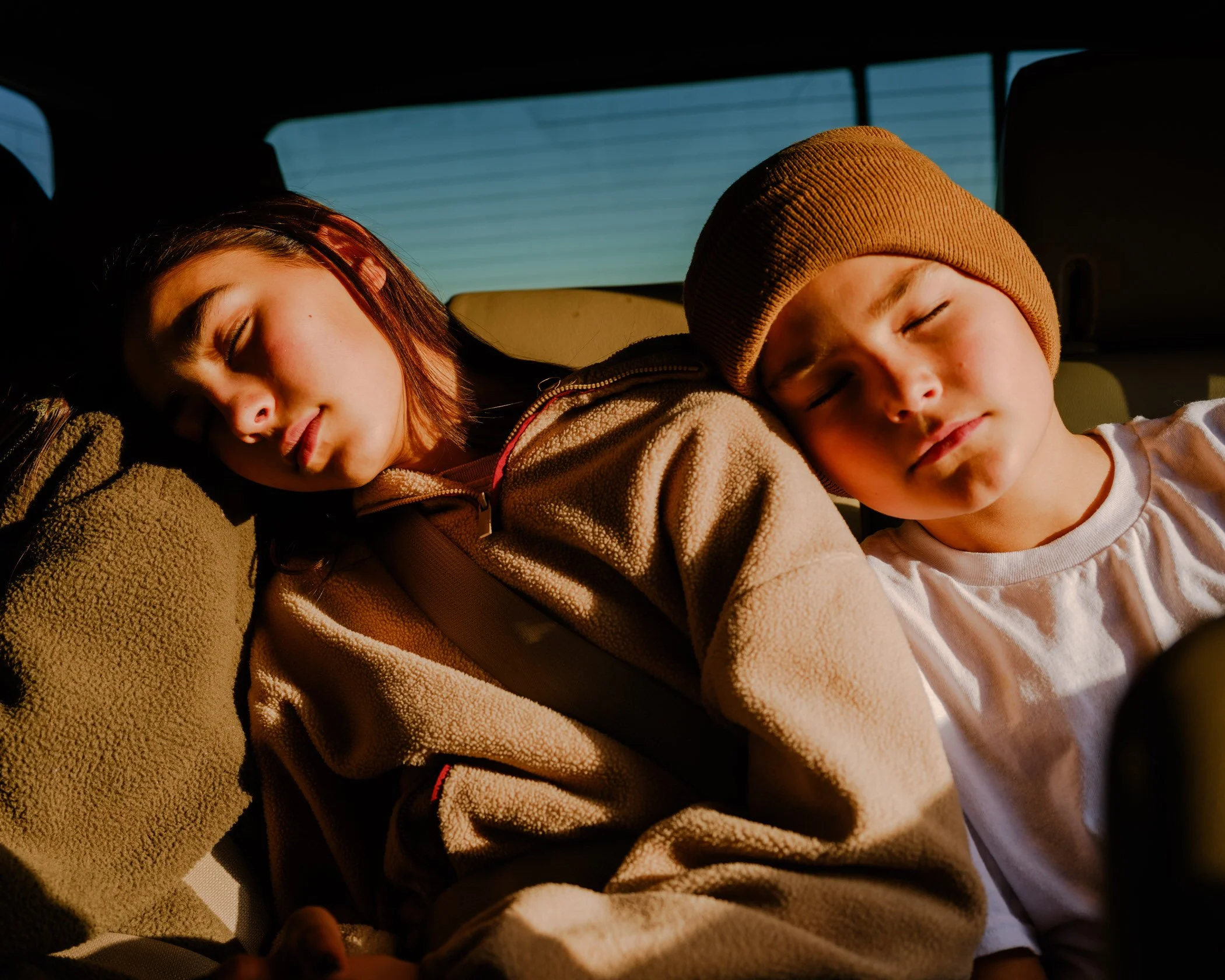 A girl and boy sleeping in the backseat of a car with sunlight shining on their faces.