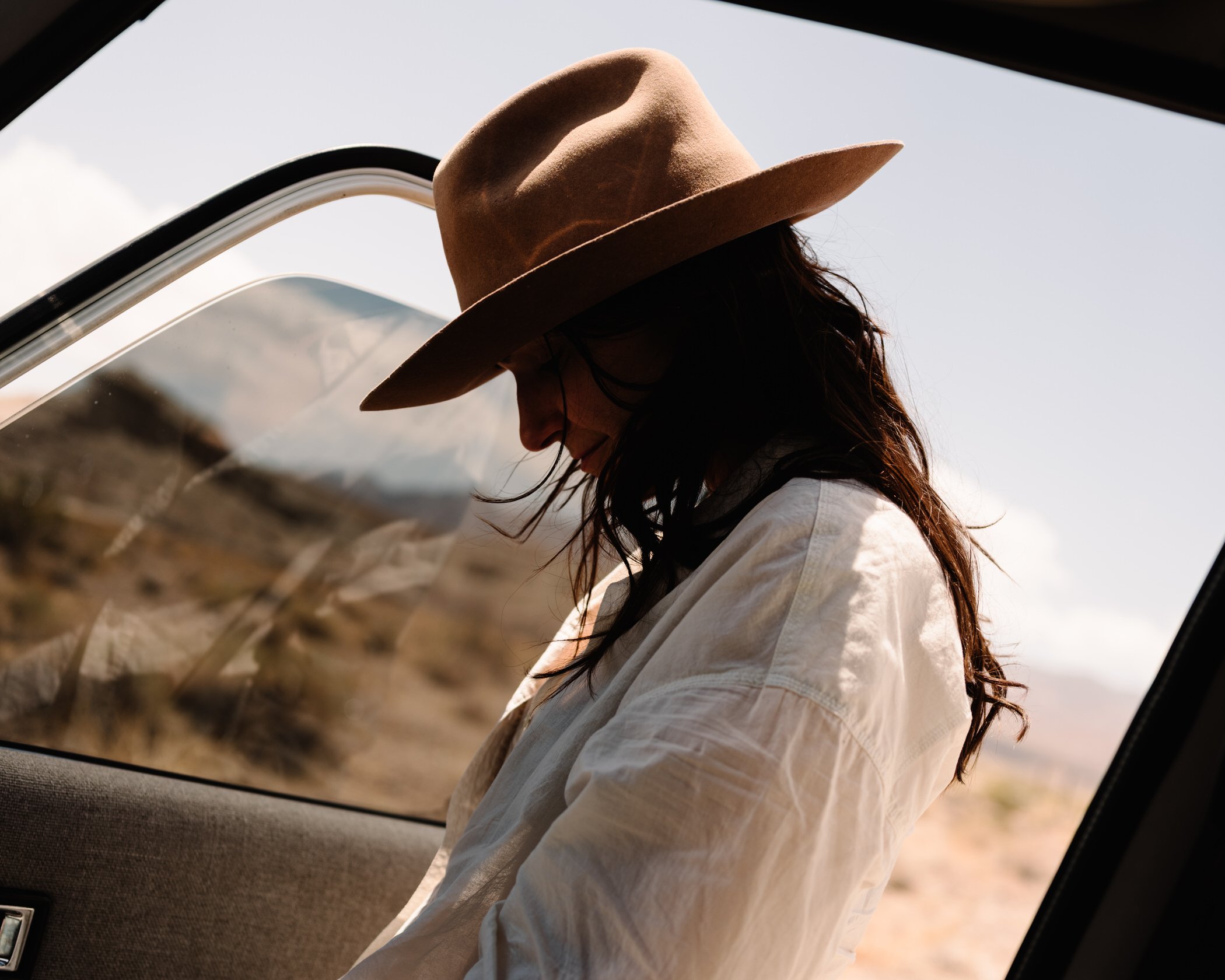 A person with long dark hair wearing a tan wide-brimmed hat and a light-colored shirt, leaning against a car window outdoors during daylight.