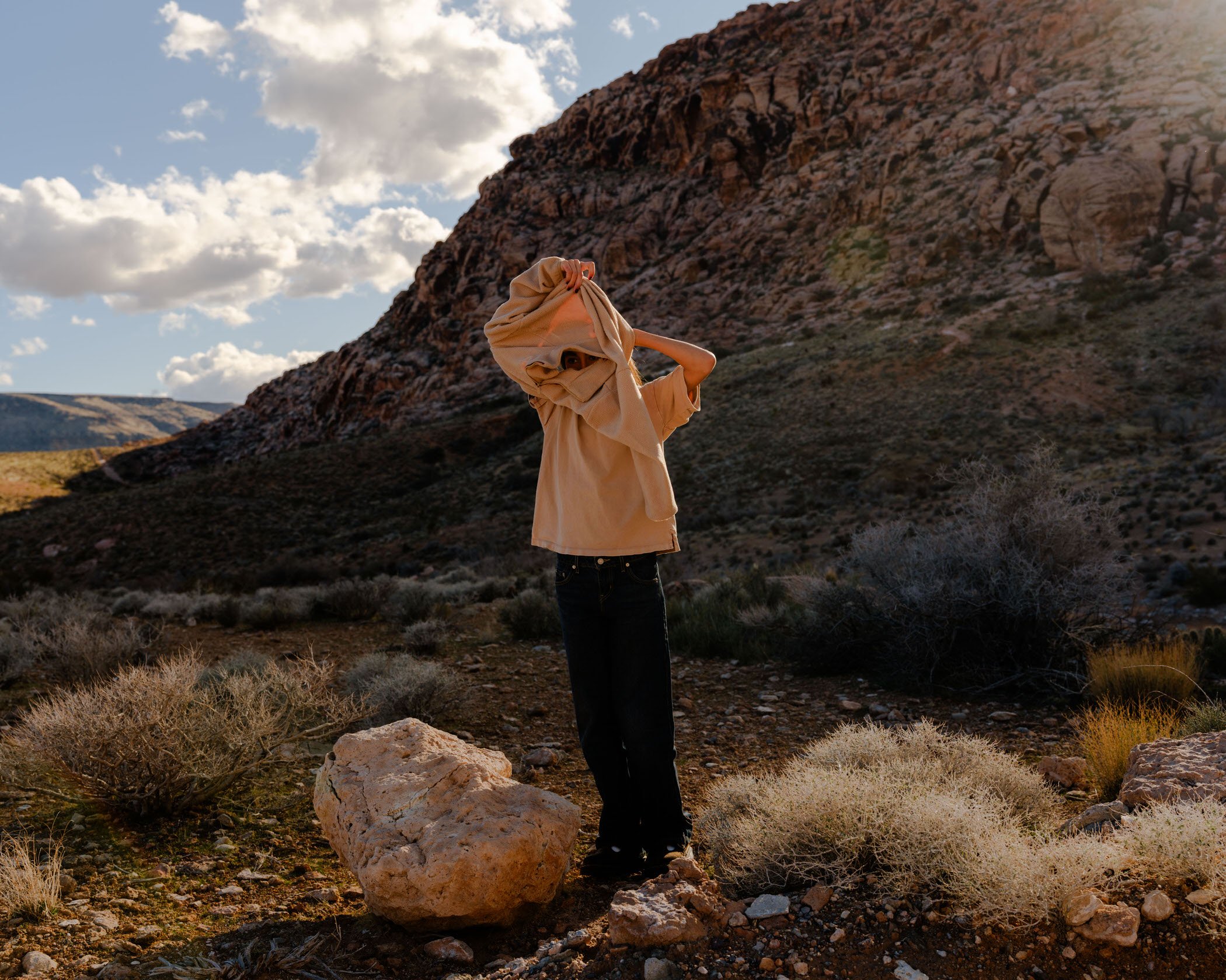 A person with dark hair wearing a beige hoodie and black pants standing on rocky terrain in front of a large rocky hill or mountain and partly covering their face with the hoodie in a desert landscape under a partly cloudy sky.